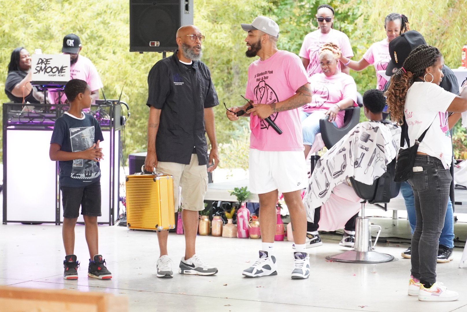 A diverse group of people gathered at an outdoor event, with some sitting and some standing. Two men are having a conversation in the center while others are engaged or observing. There is equipment and decorations nearby, and a DJ in the background.