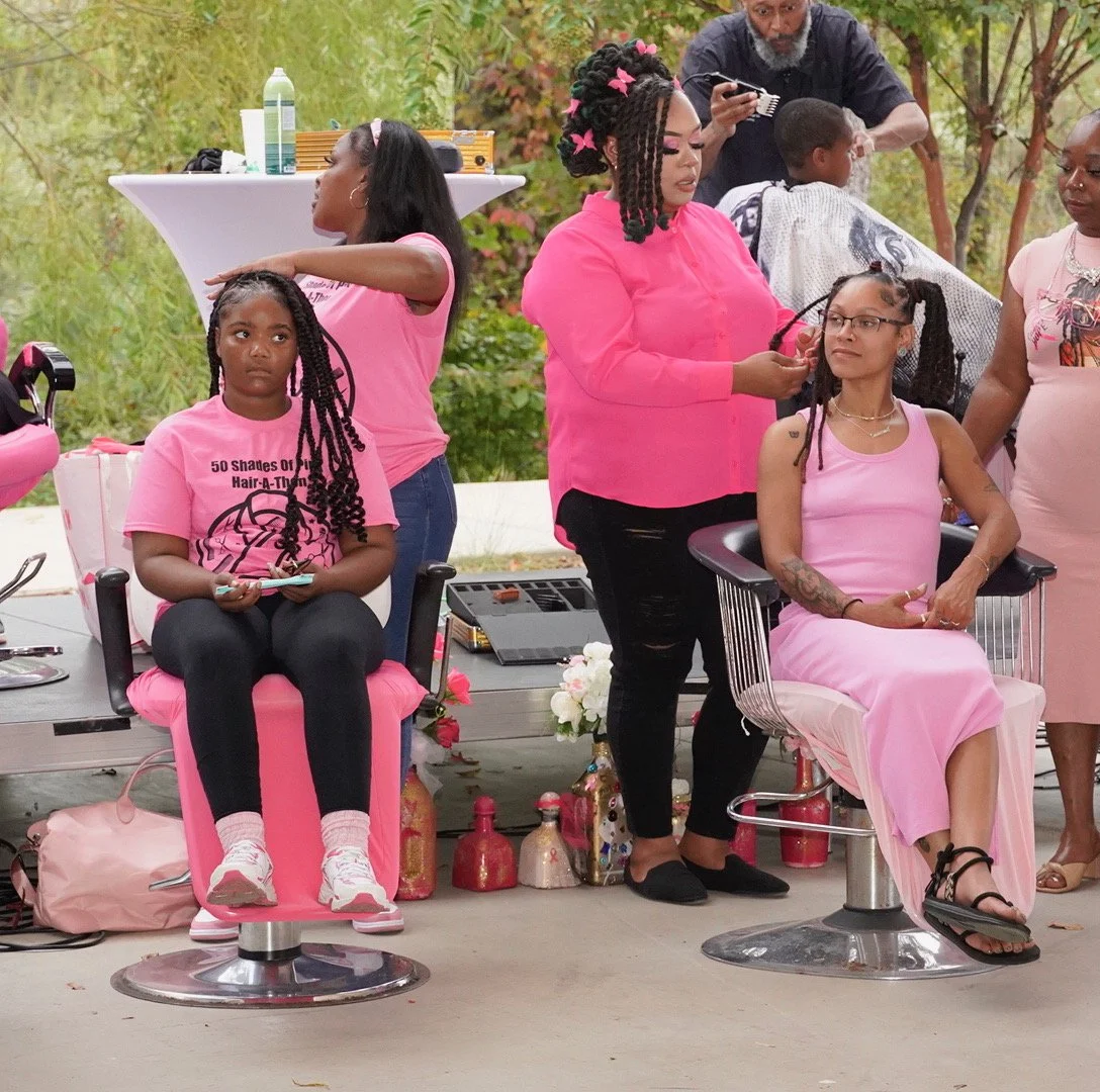 A group of women and children at a community hair event outside, with hairstyling equipment and supplies, some women getting their hair styled, and others waiting or observing.