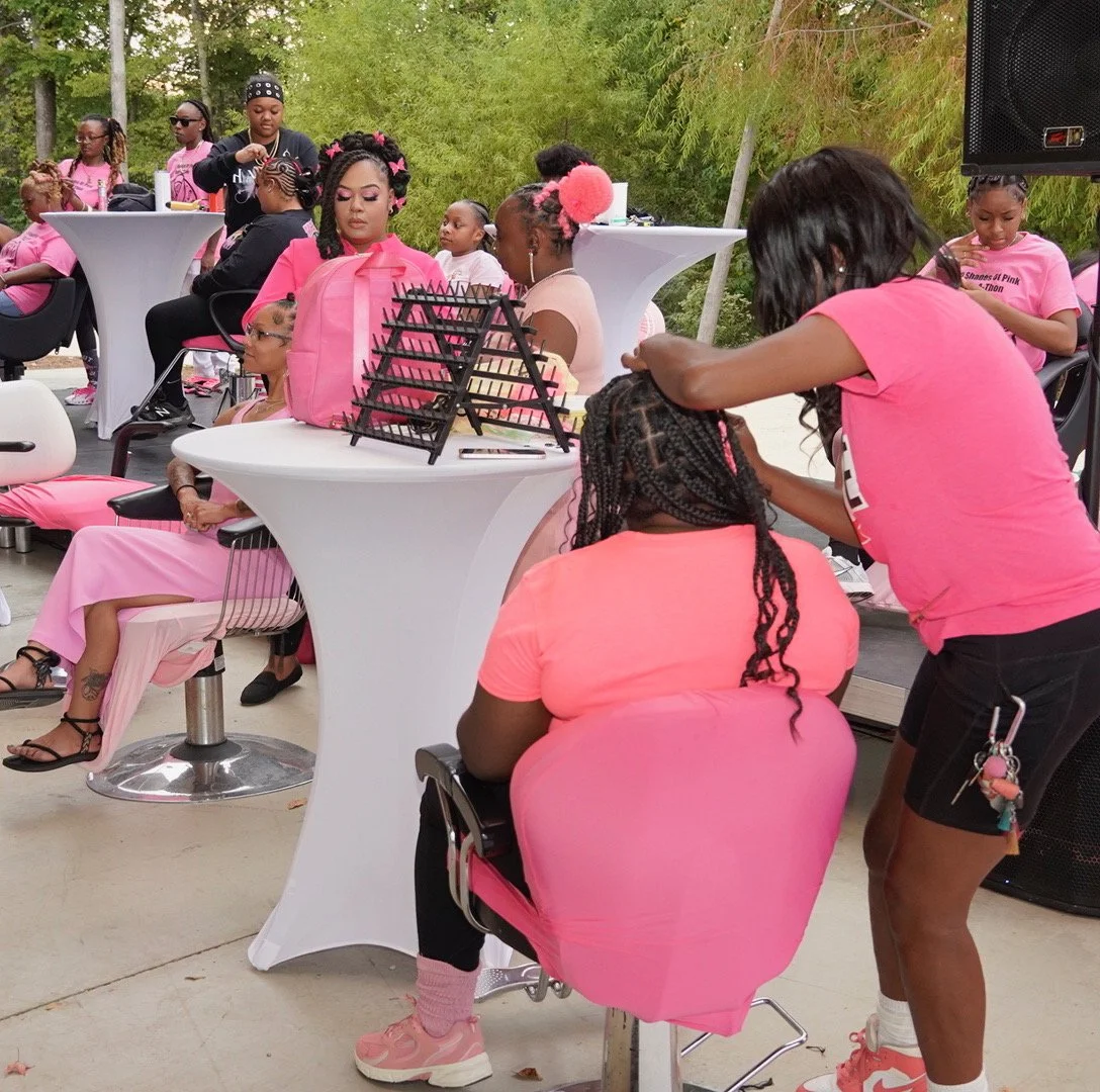 A group of women and girls gathered outdoors, dressed in pink, at a pink-themed event taking place in a park or backyard, with some sitting at small round tables and others standing, engaging in activities like getting hair styled.