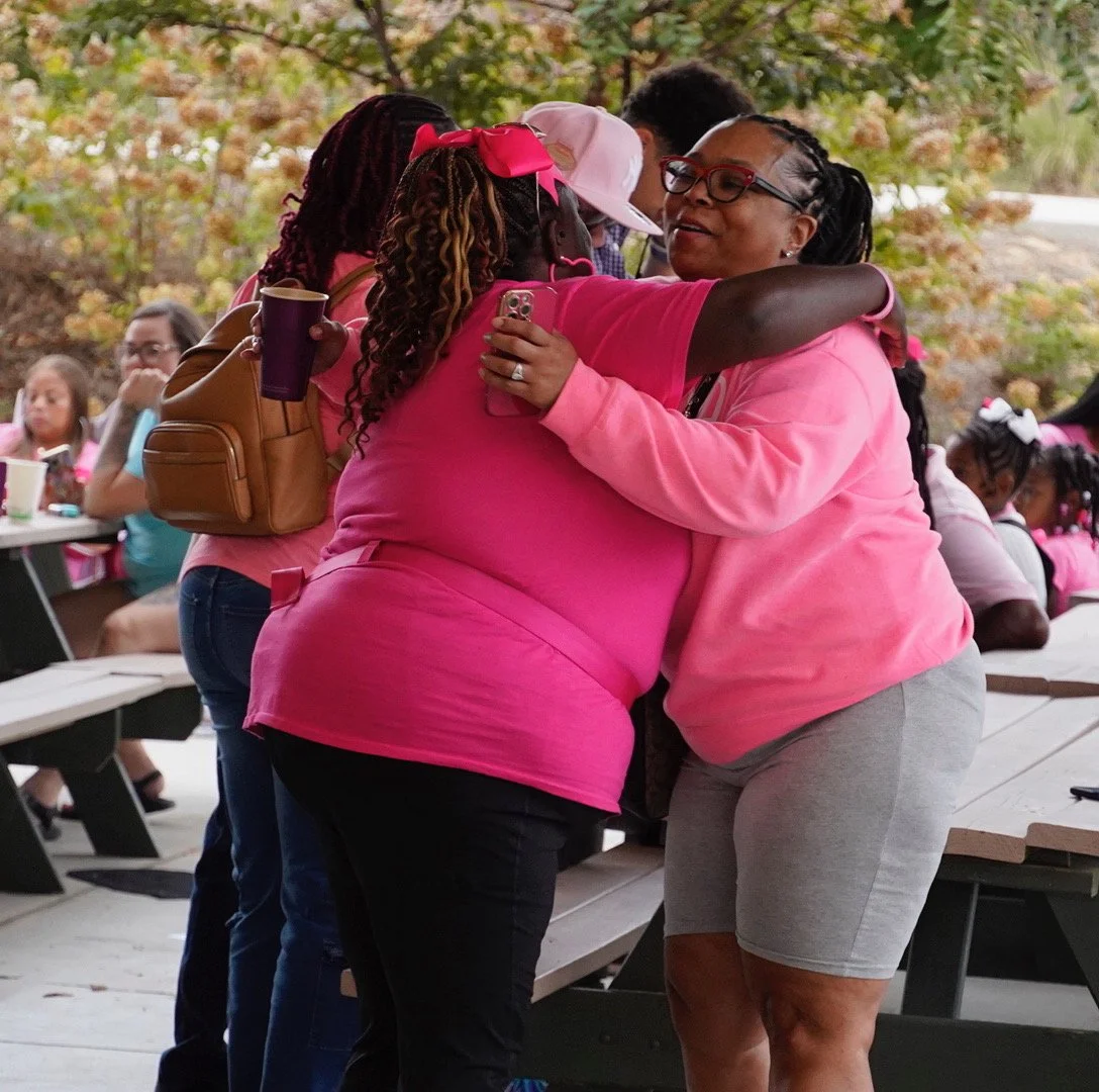 Two women hugging at an outdoor gathering, with others sitting at tables in the background.