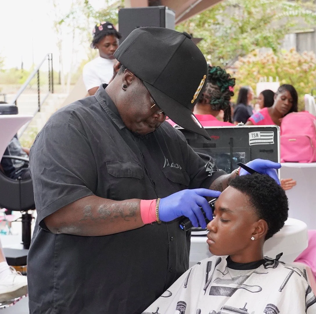 A barber wearing a black shirt, black cap, blue gloves, and glasses is shaving a woman's face at an outdoor event with people in the background.