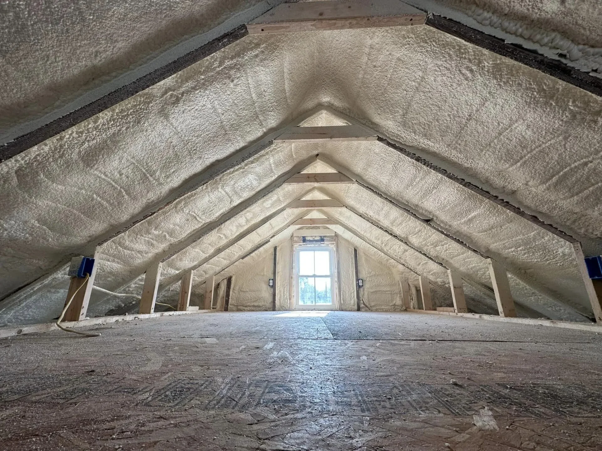 A room under construction with insulation foam sprayed in the attic space, visible wooden framing, and a window at the far end.