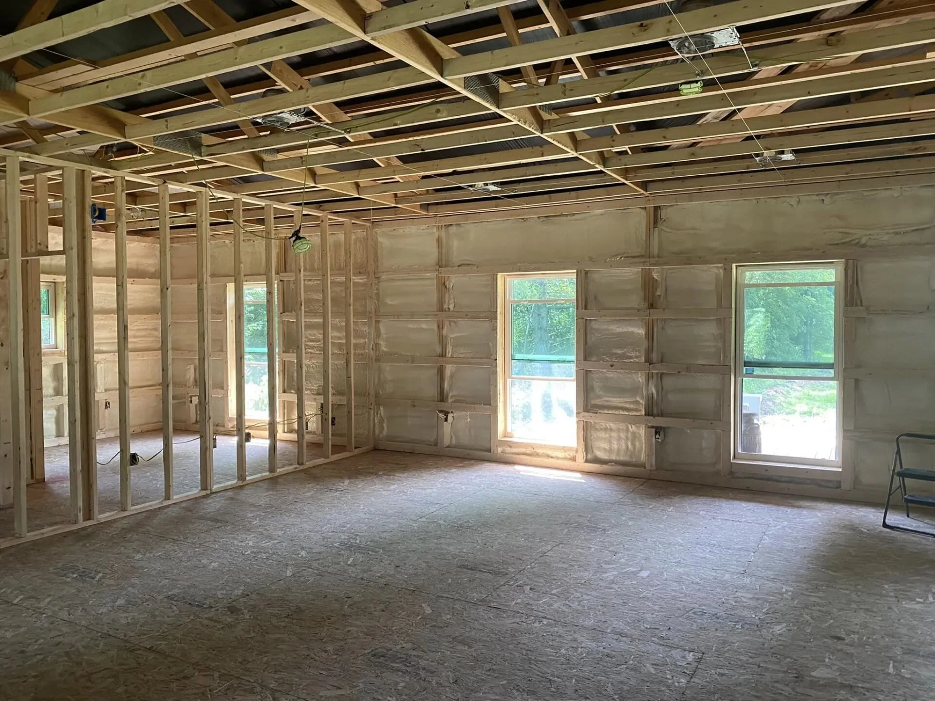 Interior of a house under construction, showing exposed wooden framing, unfinished walls, and a ceiling with open beams. Two windows are visible, with green trees outside.
