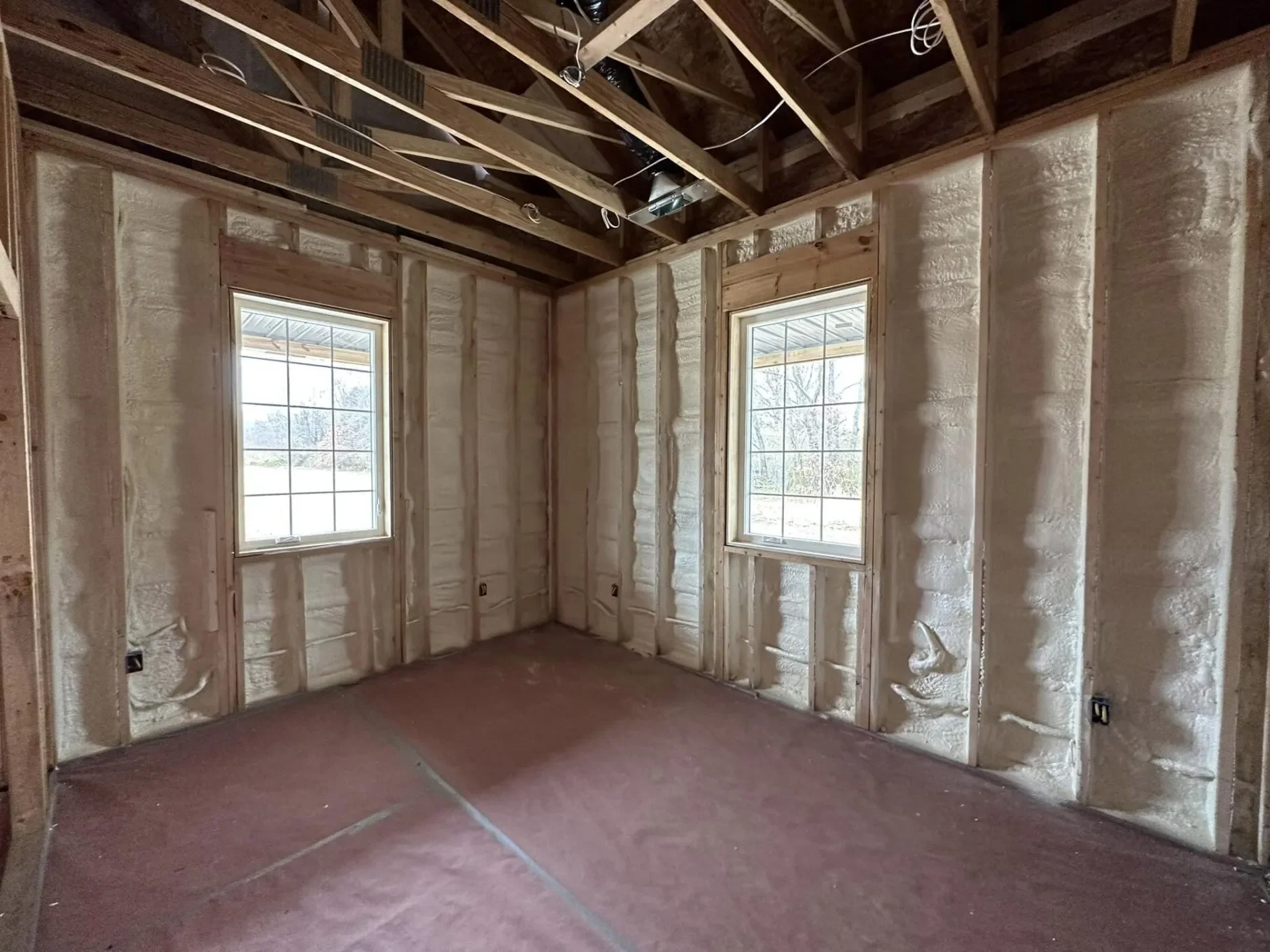 Empty room with insulation and wooden framing, two windows showing outdoor scenery, and electrical wiring in ceiling