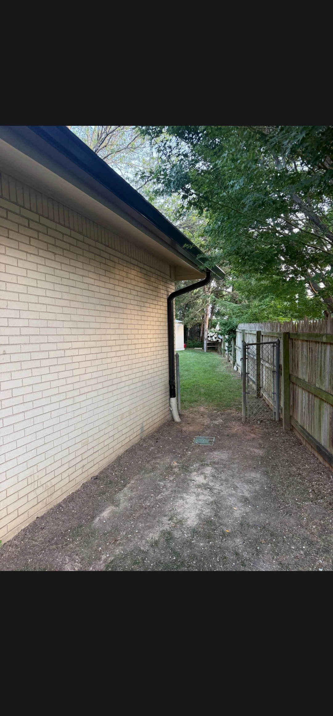 Backyard with a brick house wall on the left, a dirt area in the foreground, a grassy area with a wooden fence on the right, and trees providing shade on the right and background.