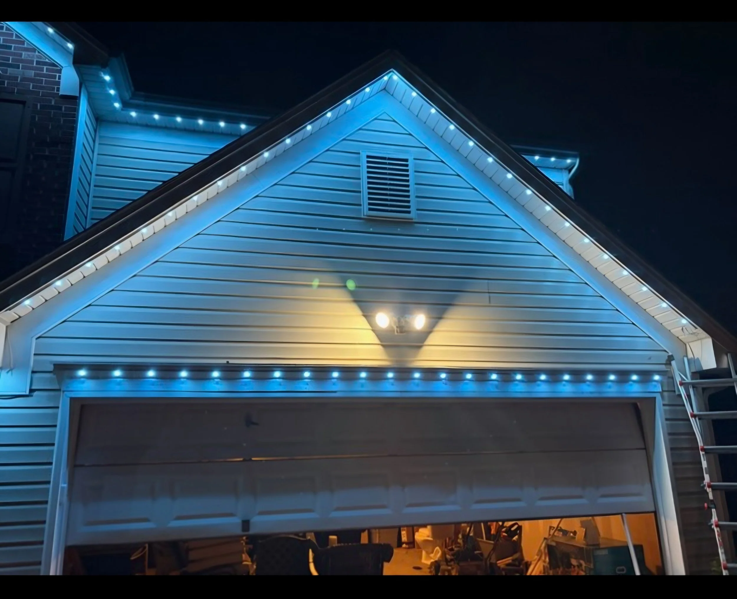 Nighttime photo of a house's front gable illuminated with white and blue LED string lights, with a vent in the center of the gable.