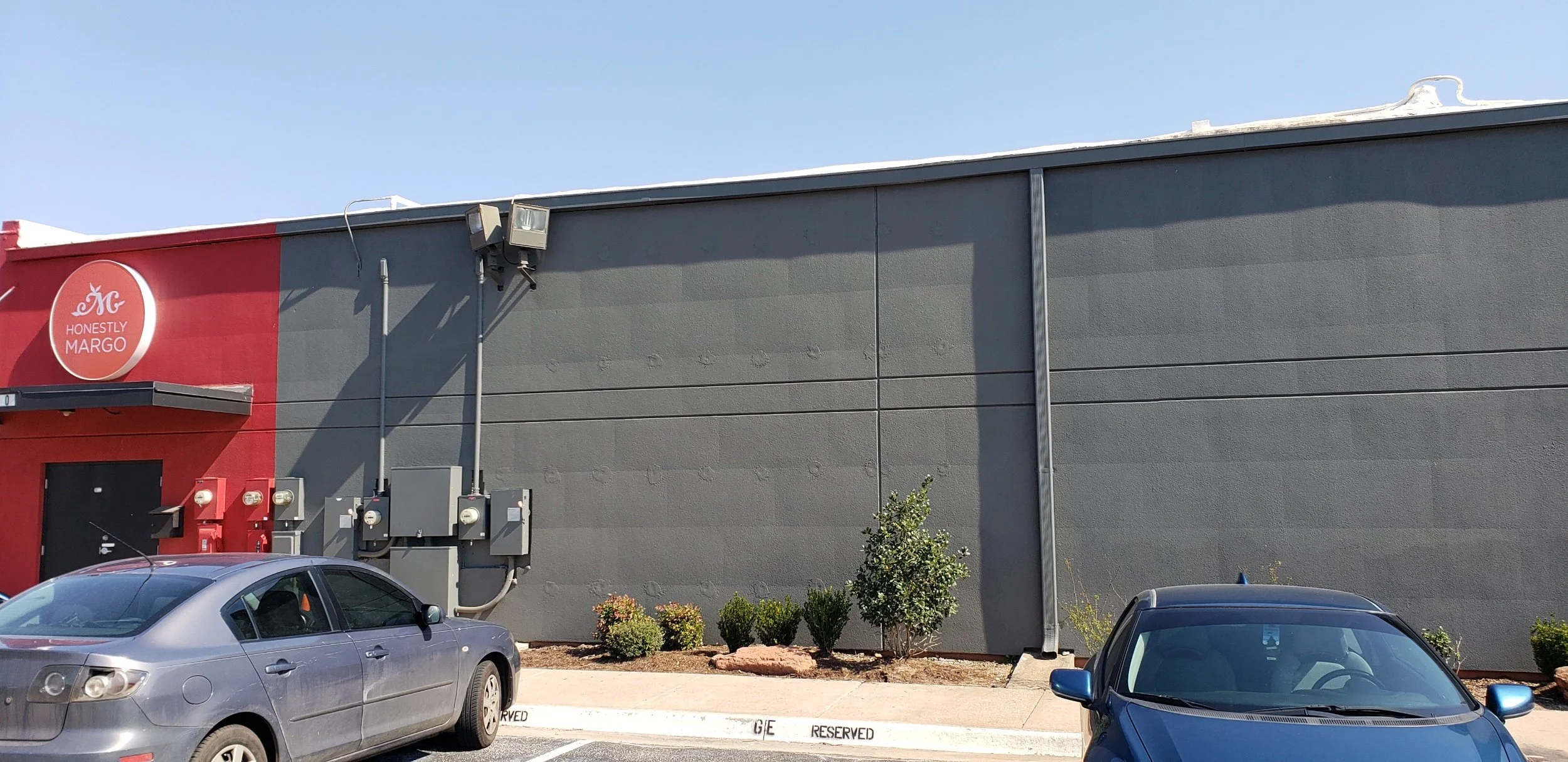 The side of a commercial building with a gray wall and a red section displaying a round logo with the words 'Honestly Margo.' There are utility boxes and a vent on the wall. The parking lot in front has two cars and small shrubs along the curb.