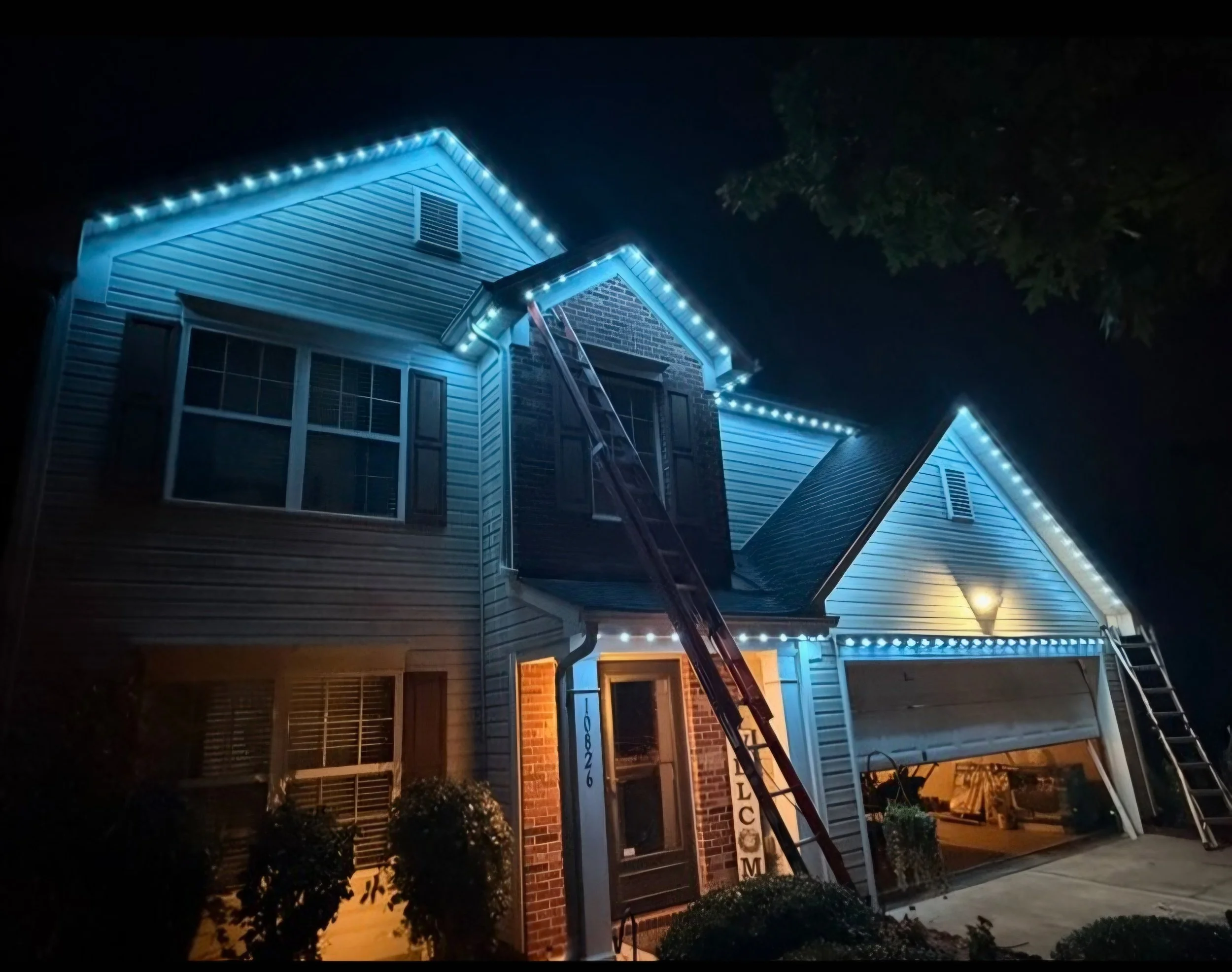 A house decorated with blue and white Christmas lights along the roofline at night, with a ladder leaning against it, and shrubs in the front yard.