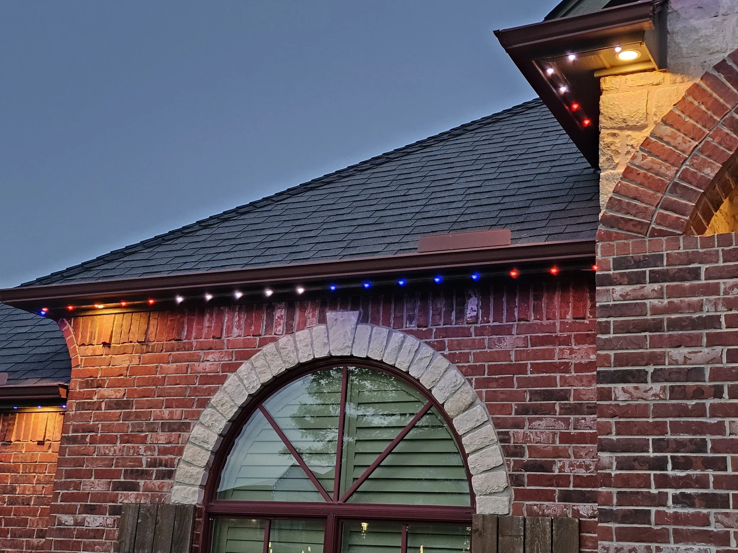 A section of a brick house with a arched window, a slate roof, and Christmas lights along the eaves at dusk.