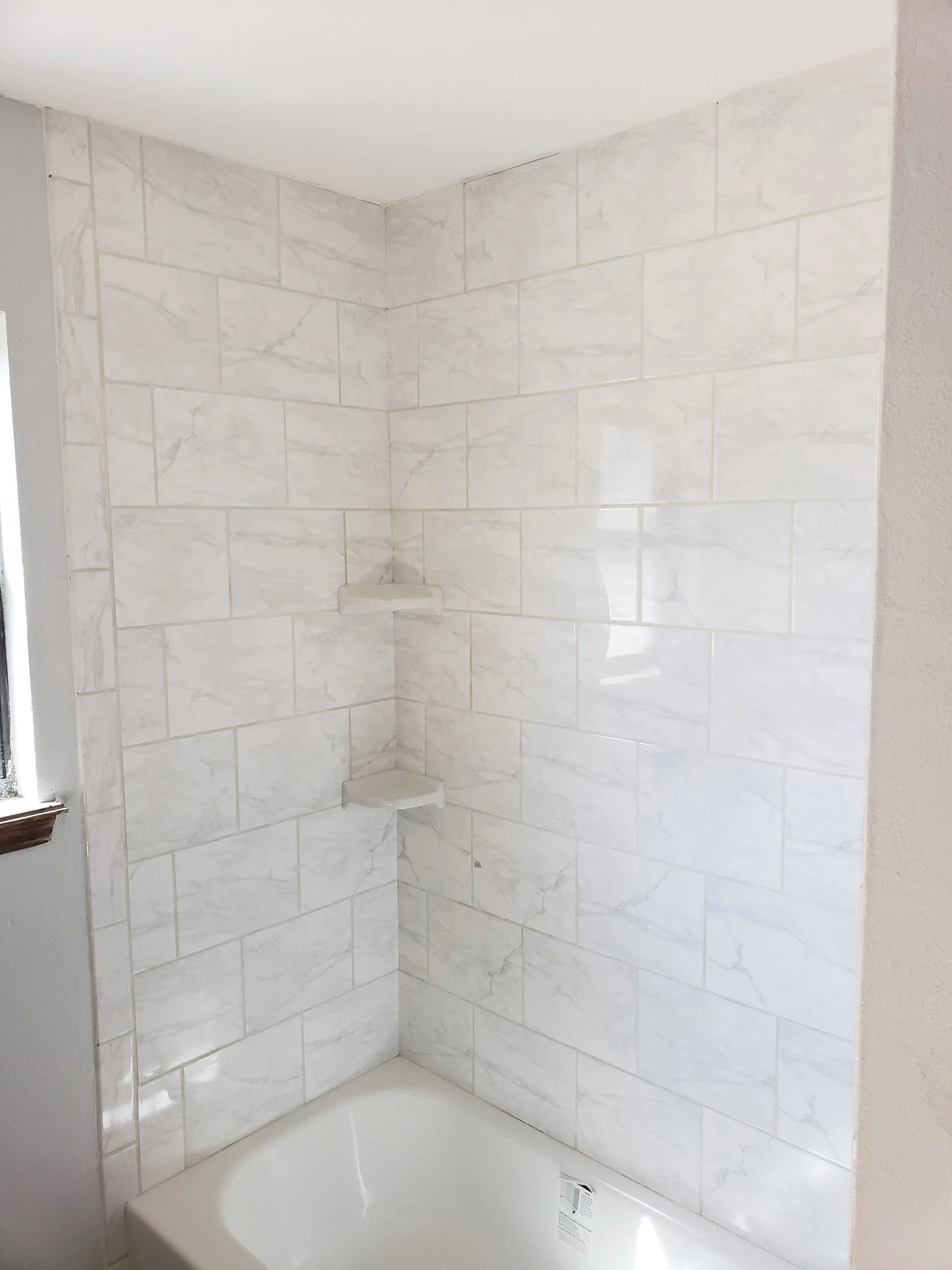 A tiled shower area with white marble-looking tiles, a corner shelf, and a bathtub at the bottom.