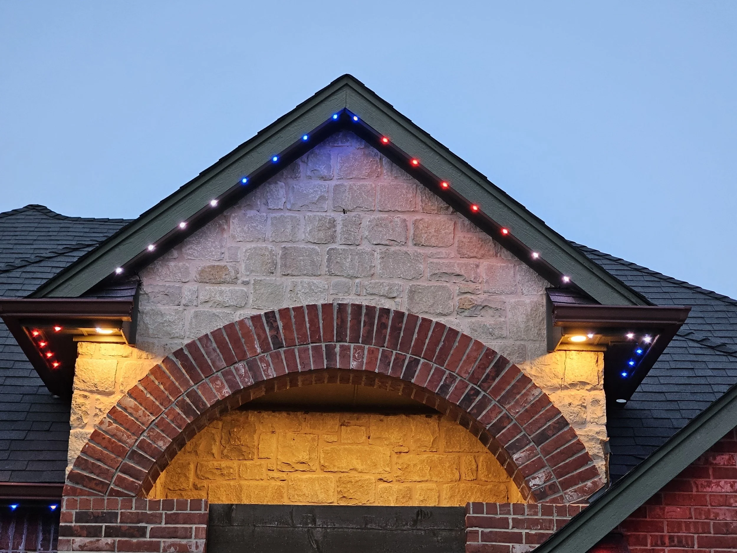 Close-up of a house's brick and stone exterior with a decorative archway. Blue and red holiday lights are draped along the roofline, and there are warm yellow lights illuminating the entrance.