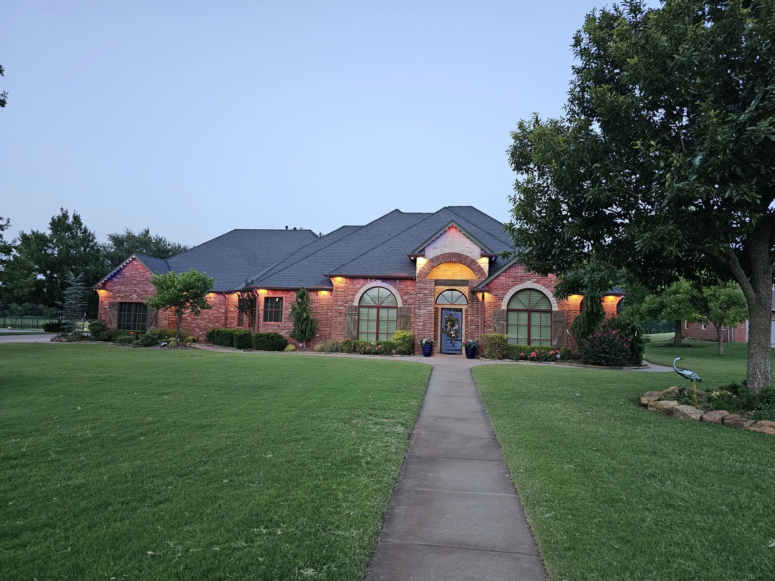 A brick house with a dark gray roof illuminated by exterior lights at dusk. The house has large arched windows and a front door decorated with a wreath. The front yard features a walkway leading to the house, green grass, trees, and shrubs, with a de