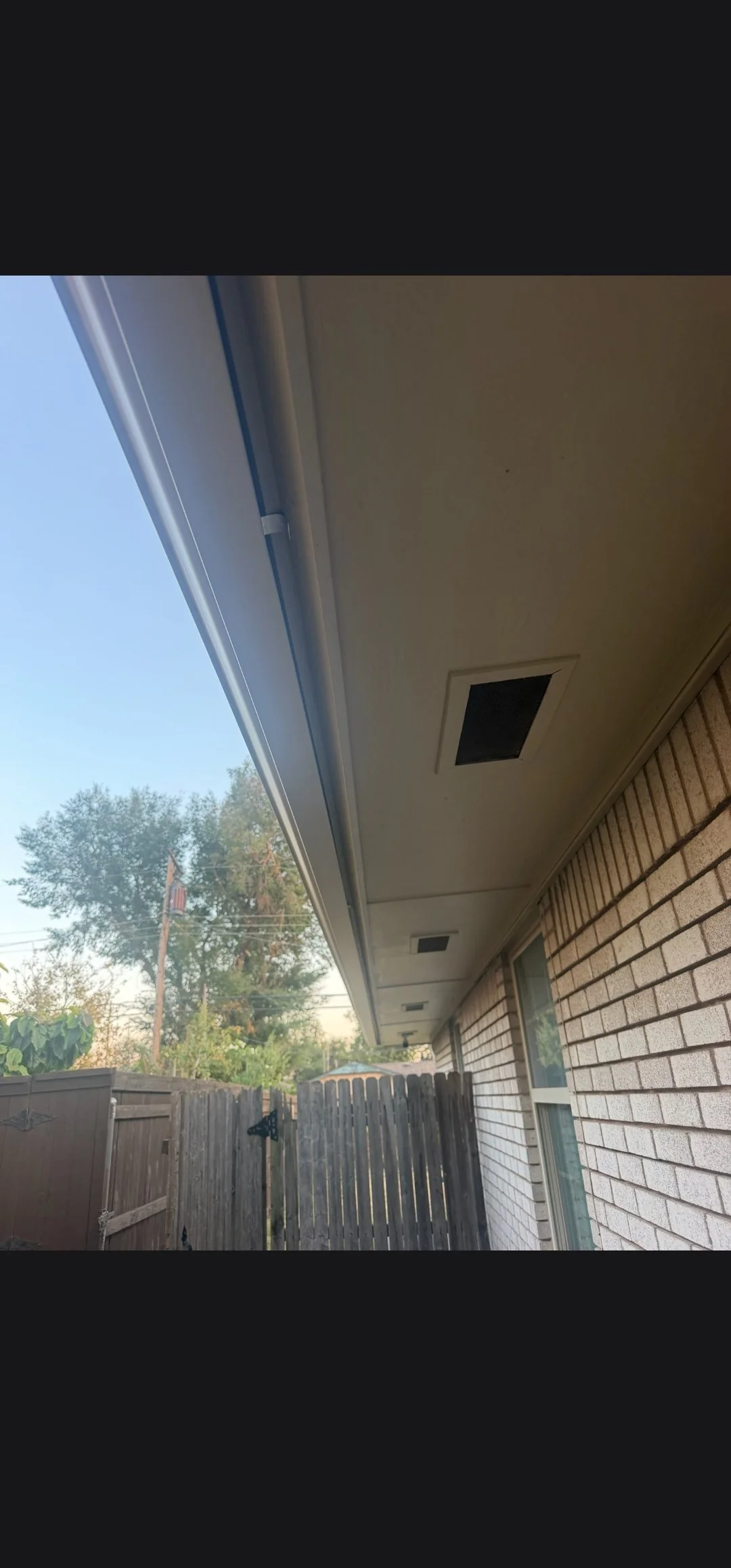 View of the back porch and backyard of a house with a brick exterior, fence, trees, and clear sky.