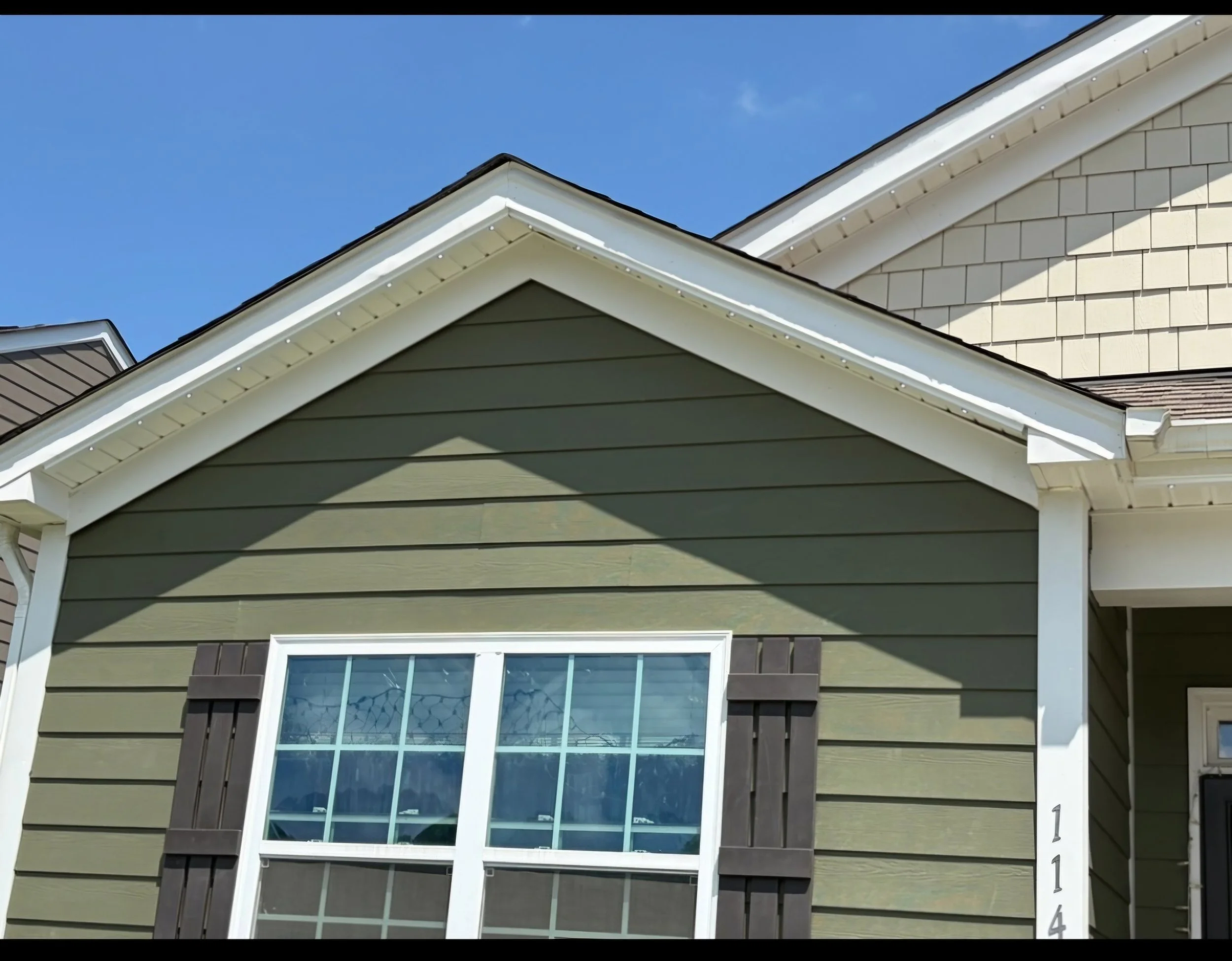Close-up of the upper part of a house showing a large window with multiple panes, green siding, white trim, and a dormer window with beige shingles on the roof against a clear blue sky.