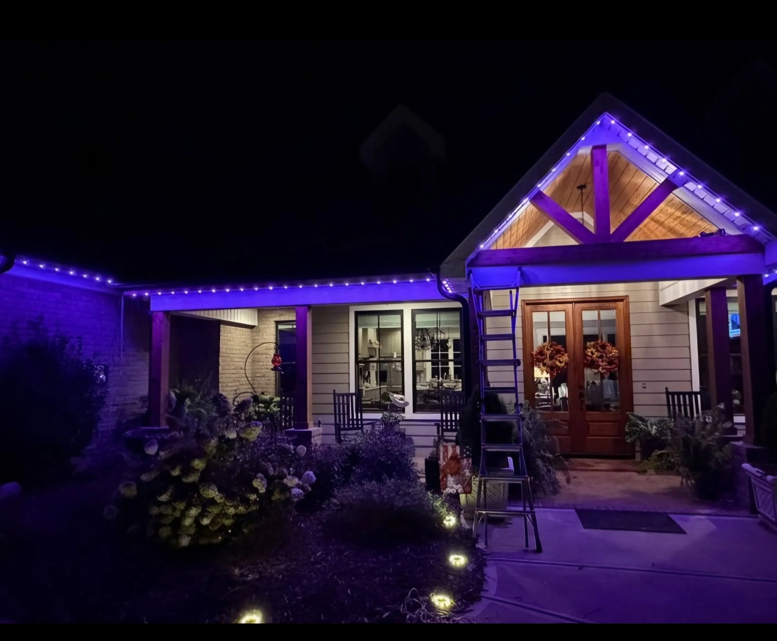 A house decorated with purple and white string lights at night, featuring a wooden roof and a garden with blooming white flowers and small landscape lights.