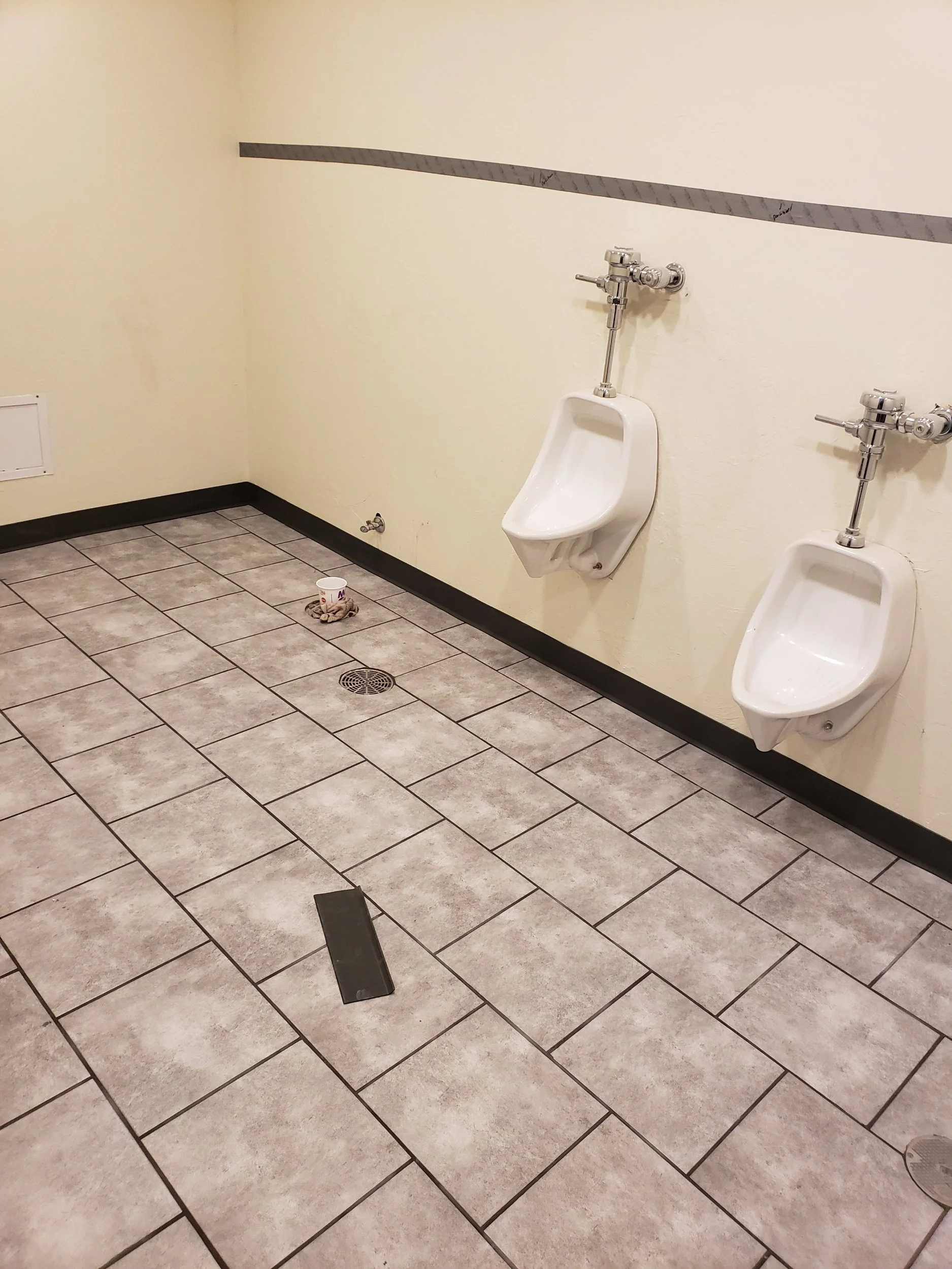 Public men's restroom with two urinals on the wall, a drain in the tiled floor, a black rubber strip on the wall, a plastic cup on the floor, and a cleaning cloth near the cup.