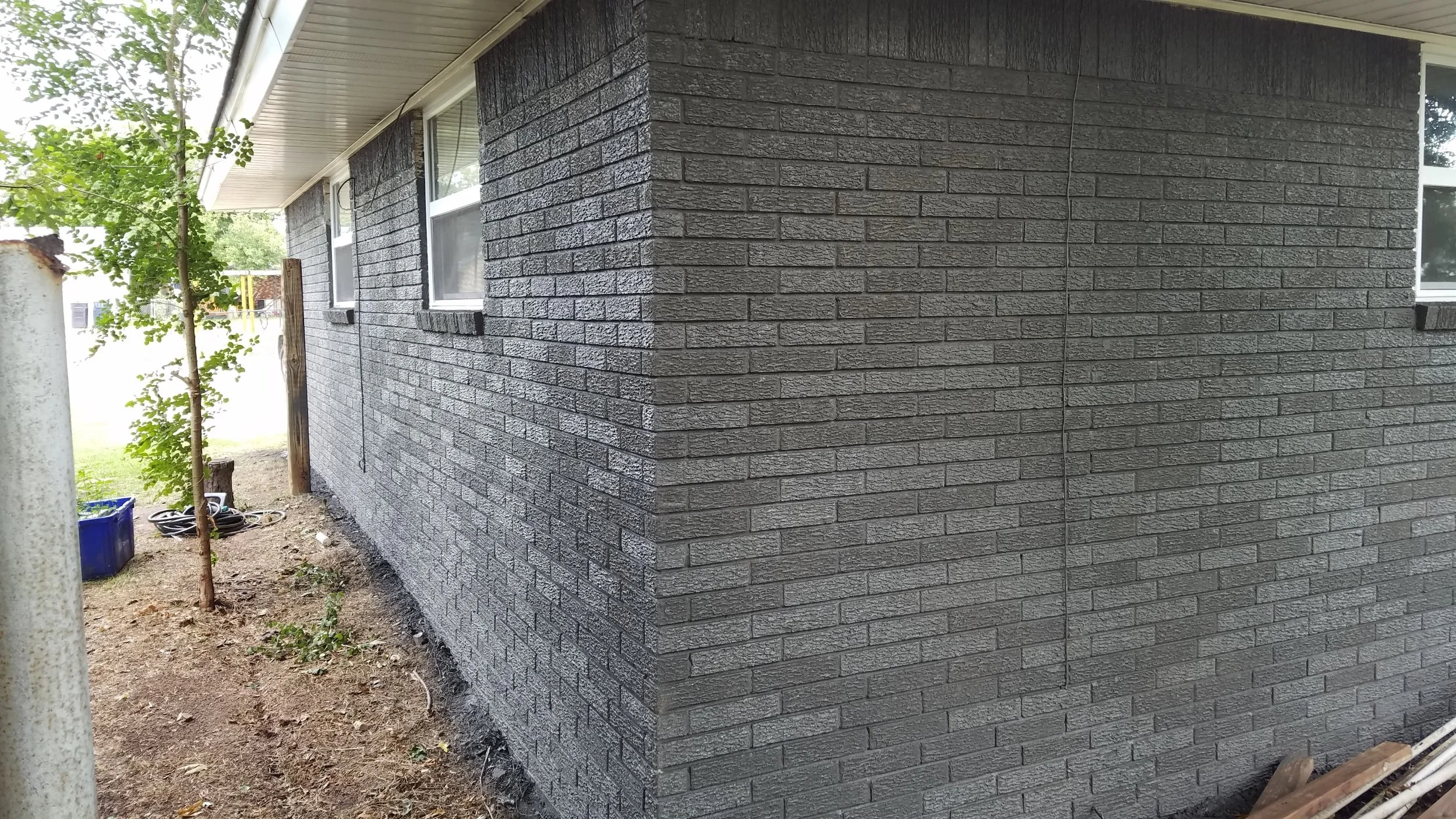 Exterior side of a house with dark gray brick siding, two small windows, and a white roof overhang. There is yard space with dirt and some gardening tools near the house, and a small tree to the left.