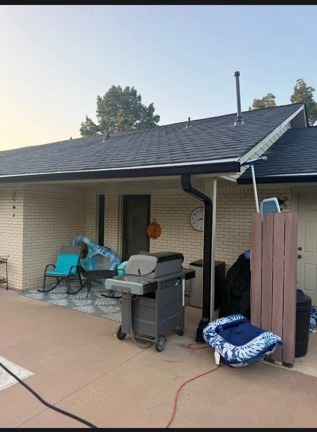 Back patio with outdoor furniture, a grill, and a small dog bed, next to a house with a brick wall and shingled roof, under a clear sky.