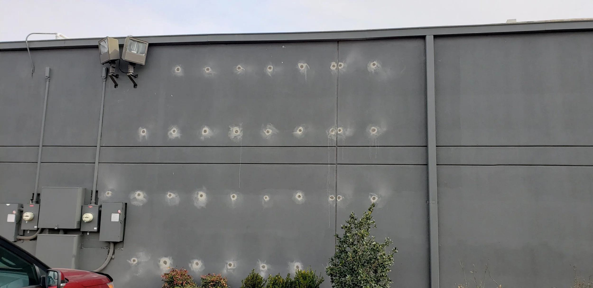 The image shows a gray exterior wall of a building with multiple visible bullet holes around the middle section. There are electrical boxes and conduits on the left side, and two outdoor lights installed near the top. A small shrub with some flowerin