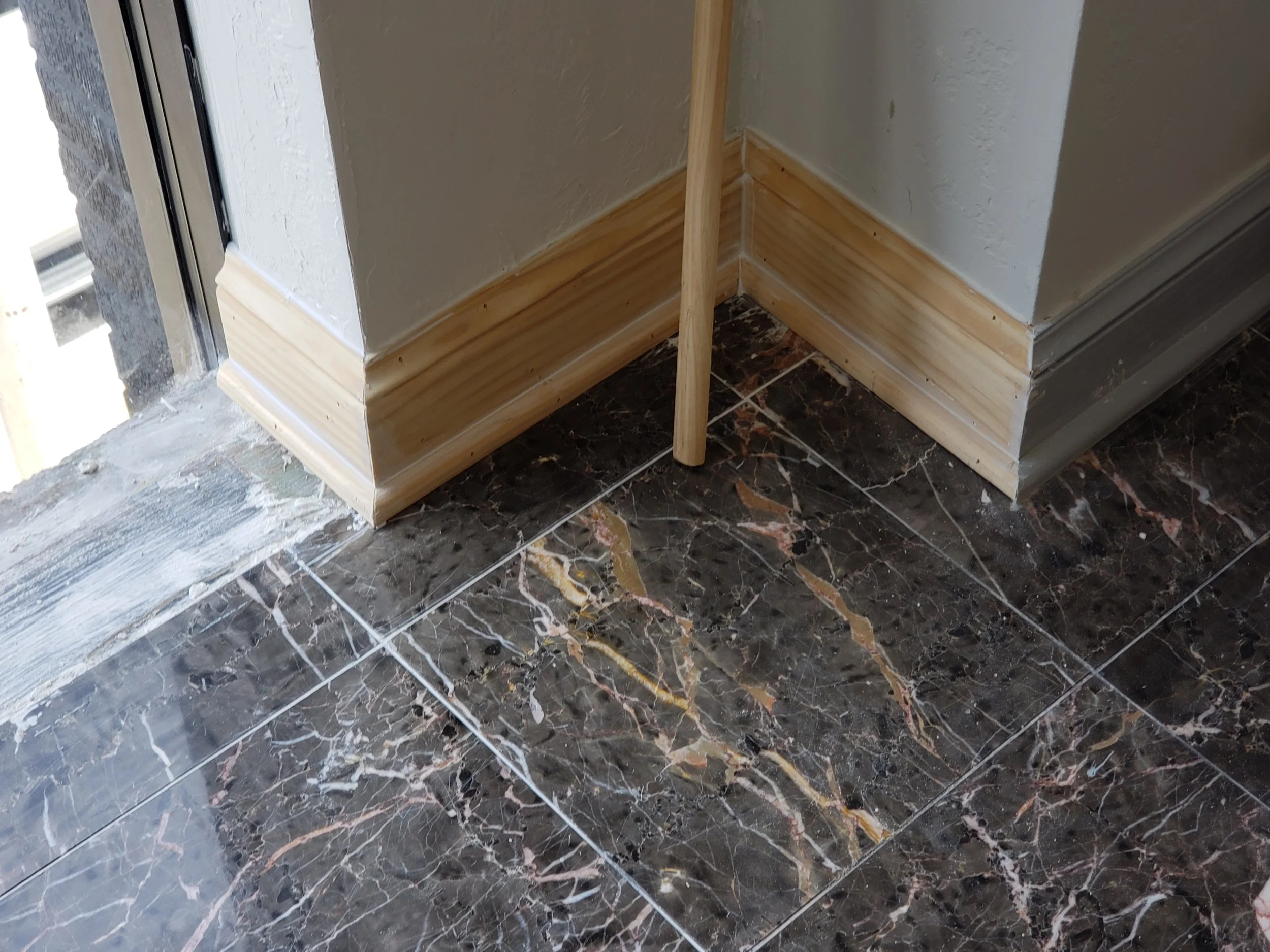 Corner of a room with marble tile flooring and wooden baseboards, next to a wall painted in a light color, with part of a sliding glass door visible on the left.
