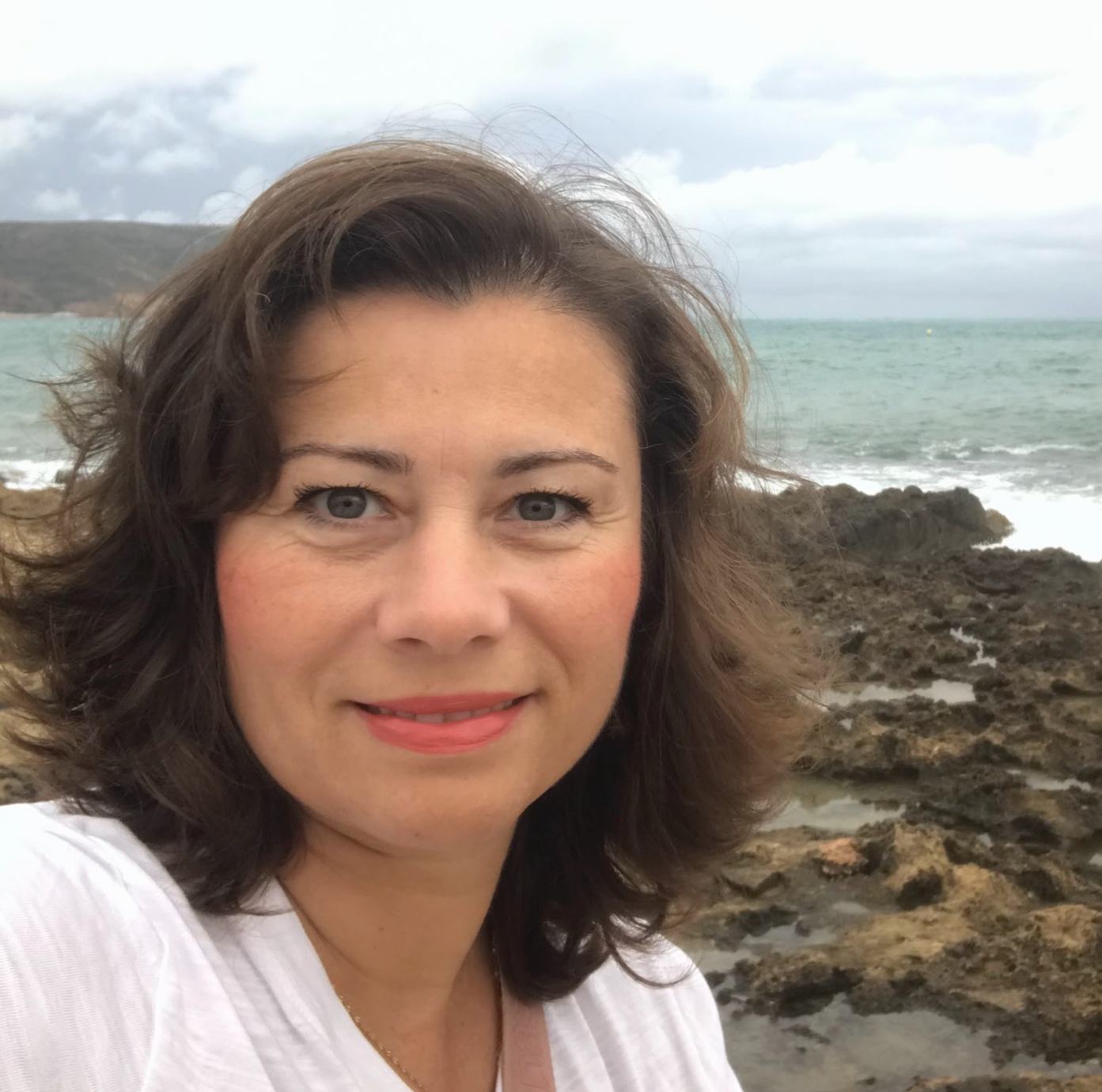 A woman with wavy brown hair and blue eyes taking a selfie at a rocky beach with the sea and cloudy sky in the background.