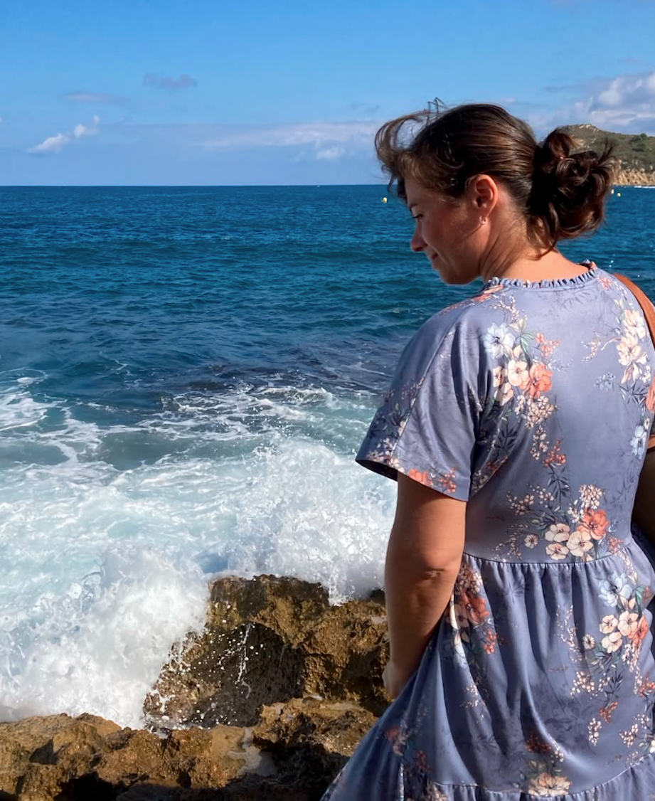 A woman in a floral dress stands on rocks by the sea, looking down at the water and waves crashing against the rocks under a blue sky with some clouds.