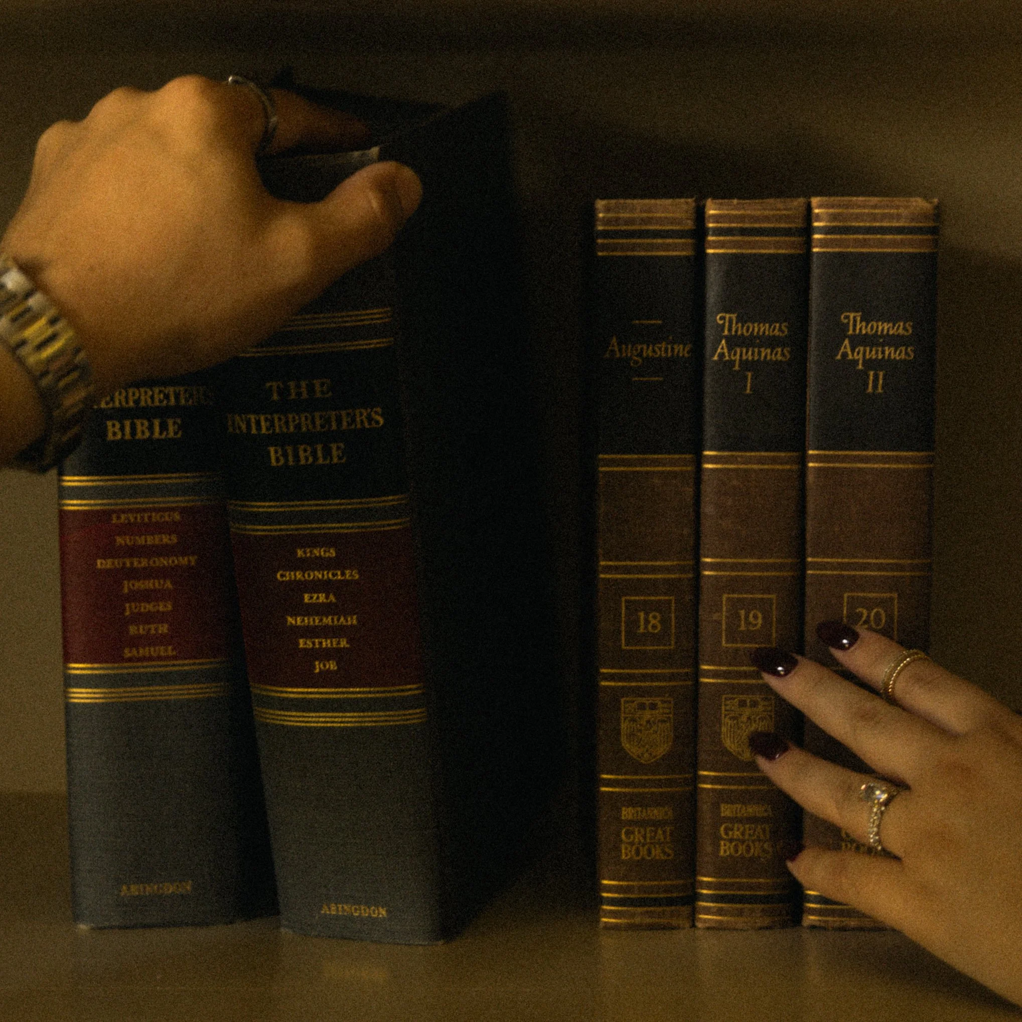 Close-up of a person's hand holding several hardcover Bible books with gold accents on the spines, including titles like "The Bible Interpreters" and "Thomas Aquinas".