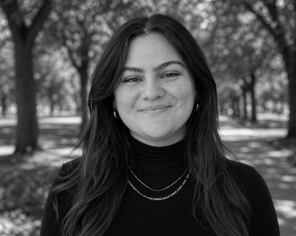Black and white portrait of a young woman with long hair, smiling, standing outdoors with trees in the background.