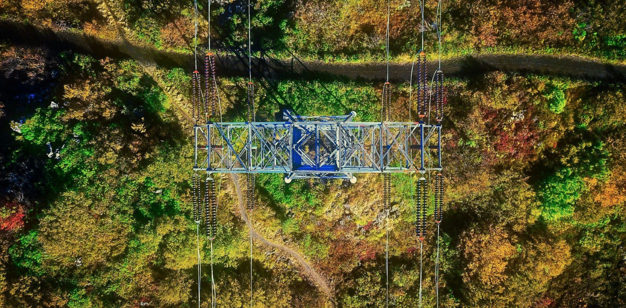 An aerial view of a transmission tower standing in a forest with colorful fall foliage.