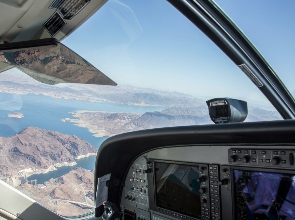 Inside the cockpit of an aircraft showing the flight instruments and controls. Out the windshield, a view of a large body of water, mountains, and land.