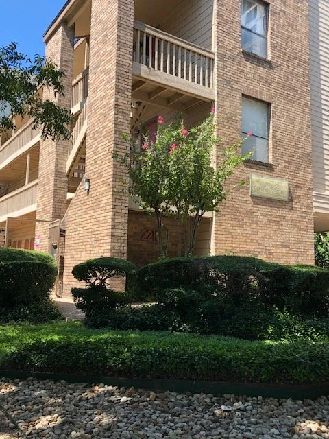 A brick apartment building with balconies, a tree with pink flowers, and neatly trimmed bushes in front.