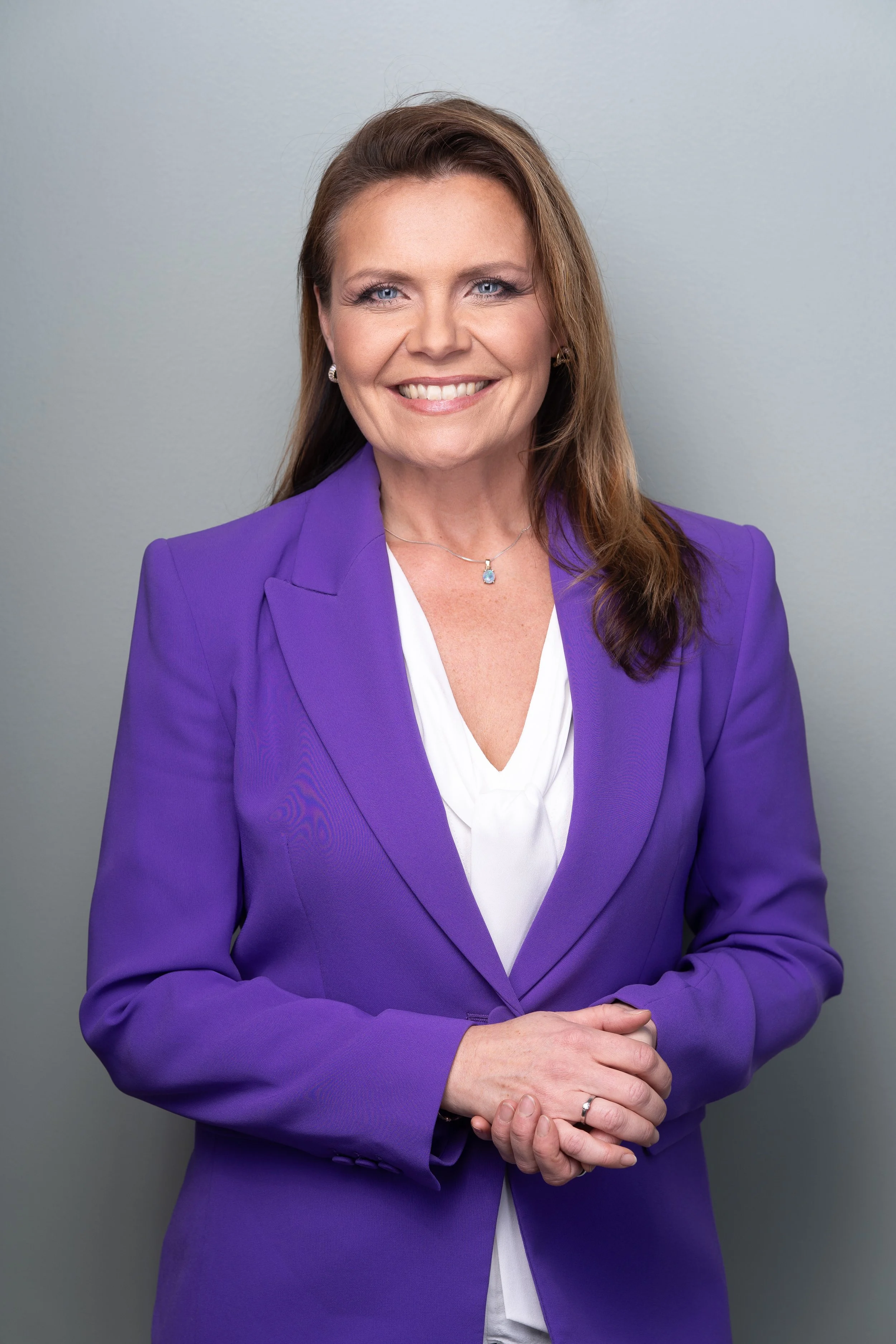 A woman with light skin color and long brown hair wearing a purple blazer, white blouse, and jewelry, smiling with her hands clasped in front of her against a plain gray background.