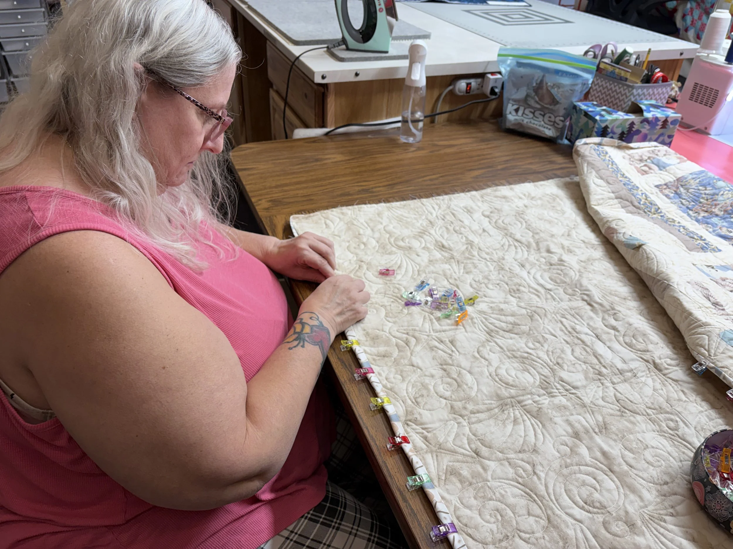 A woman with long gray hair and tattoos on her arm, wearing a pink tank top, is sitting at a wooden table working on an embroidery or quilting project. There are small colorful clips on the quilt she is working on, and sewing supplies, snack bags, and a space heater on the table.