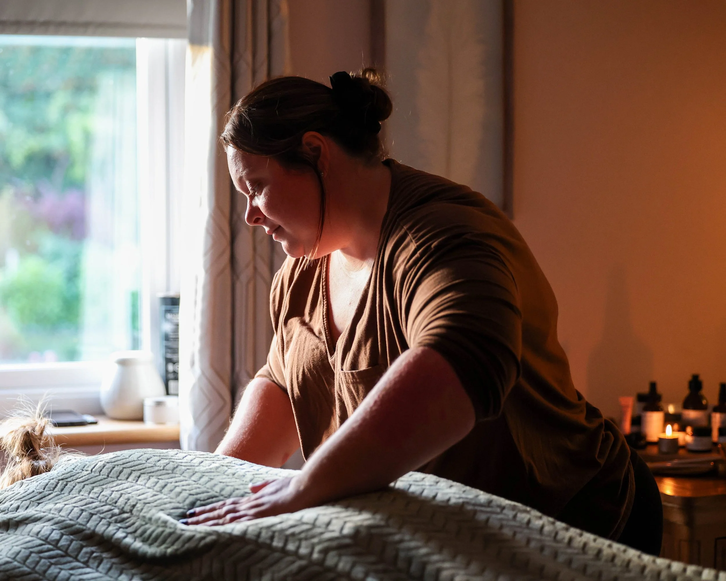 Woman leaning over a bed beside a sunlit window in a warmly lit bedroom