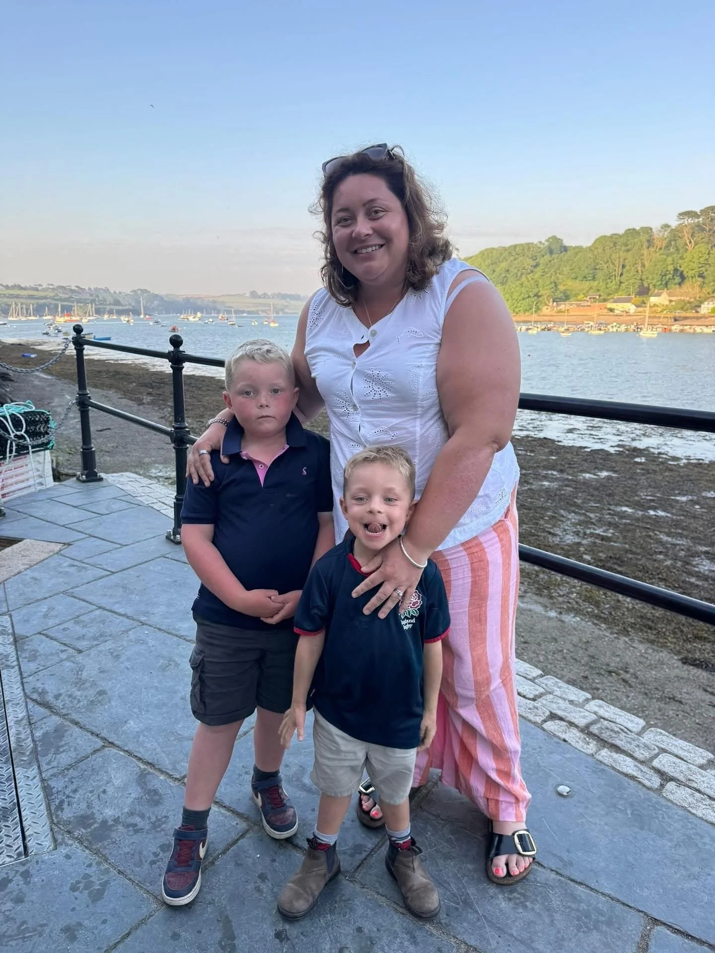 Woman posing with two young boys on a waterfront promenade overlooking sailboats and a harbor