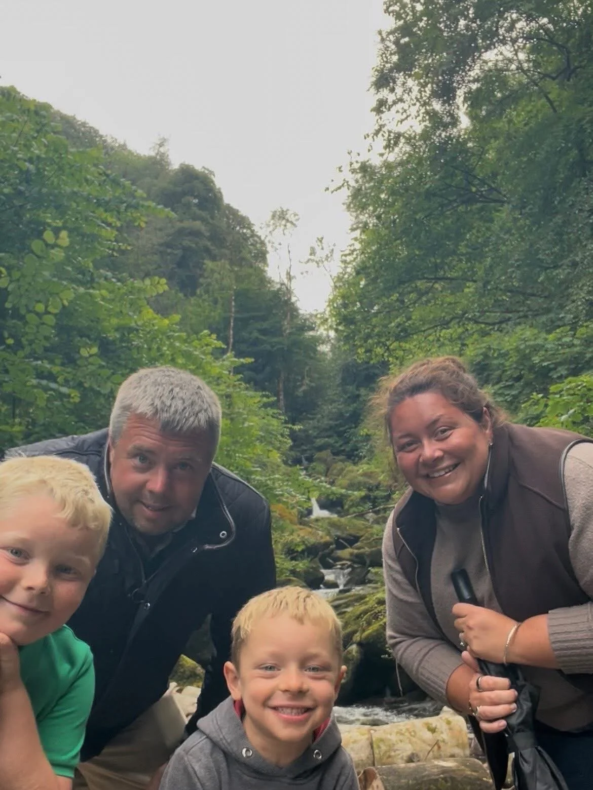 Family of four smiling beside a forest stream and small waterfall