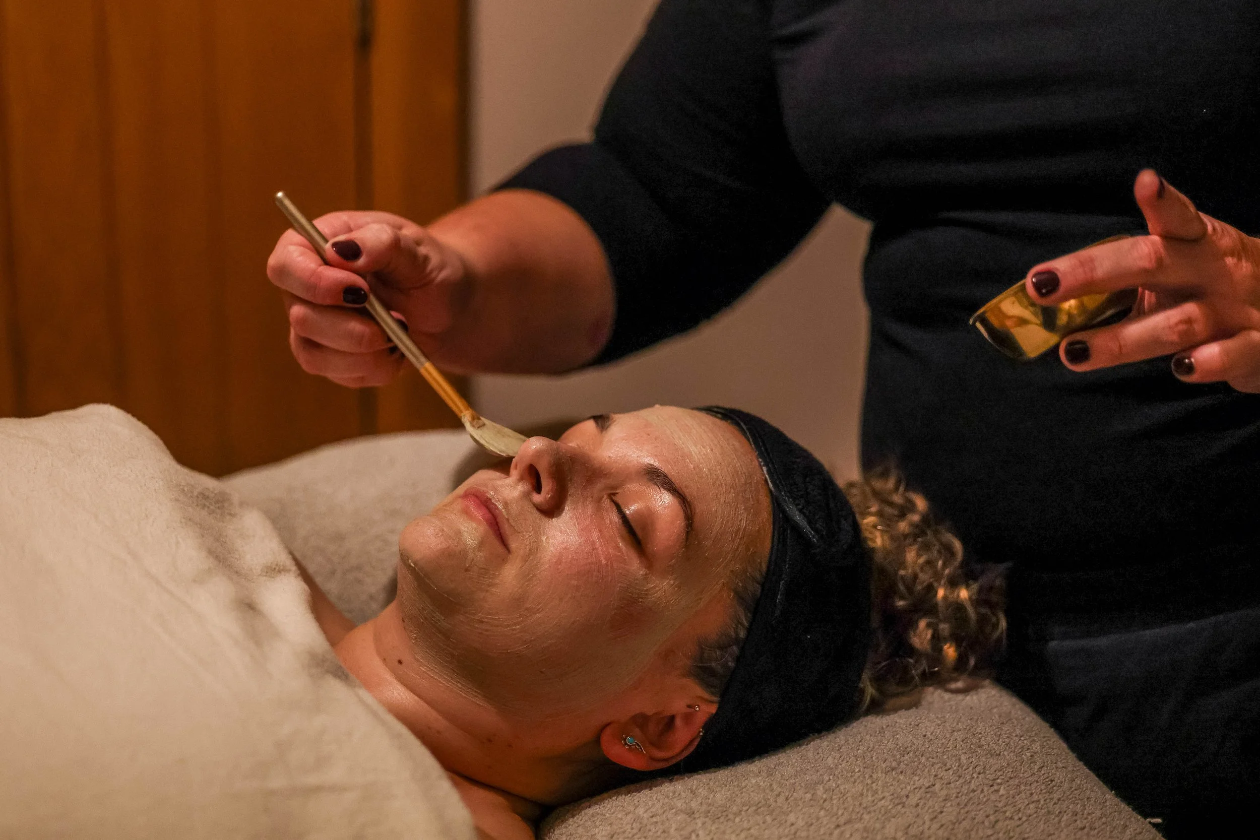 Spa aesthetician applies a facial treatment with a brush to a client lying on a treatment bed.