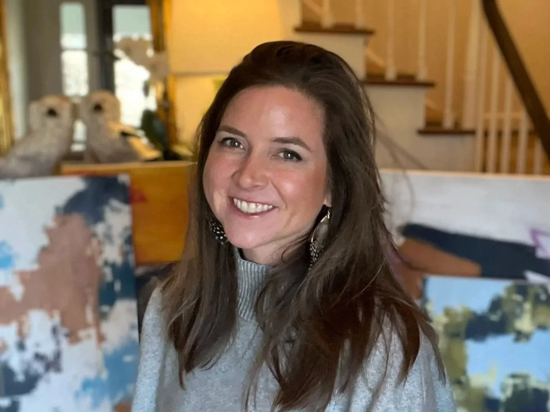 A woman with long brown hair smiling at the camera, taking a selfie in a cozy home interior.