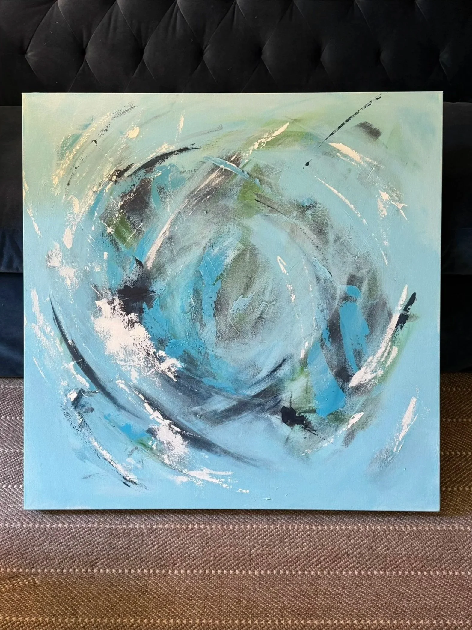 An abstract painting with swirling strokes of turquoise, blue, navy, white, and green on a light background, propped against a navy velvet sofa.