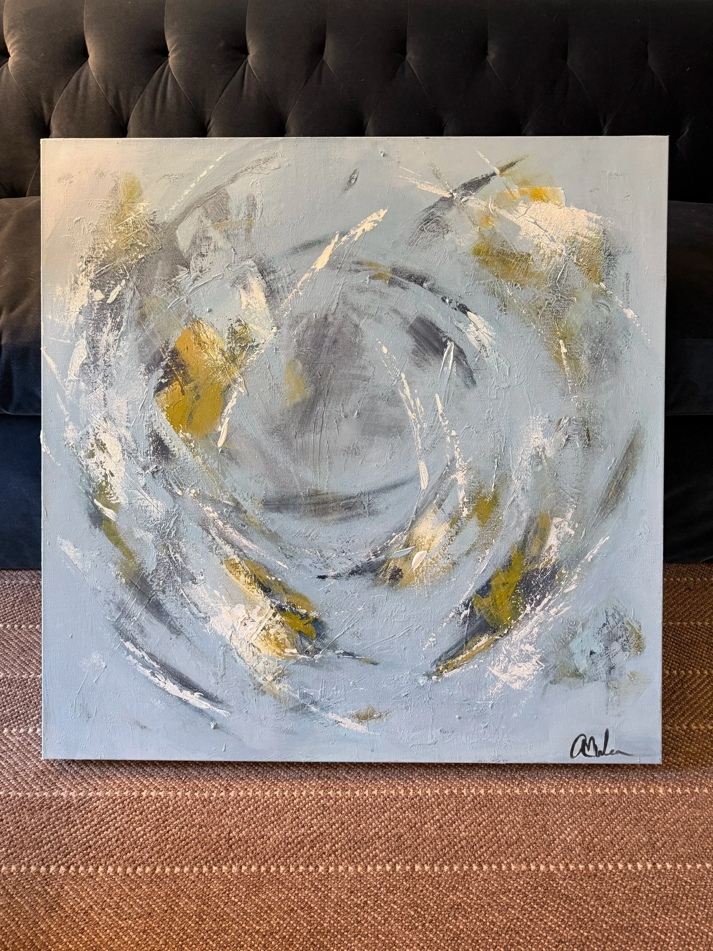 Abstract painting with swirling gray, white, navy, and gold brushstrokes, propped against a navy velvet sofa and signed by the artist in the lower right corner.