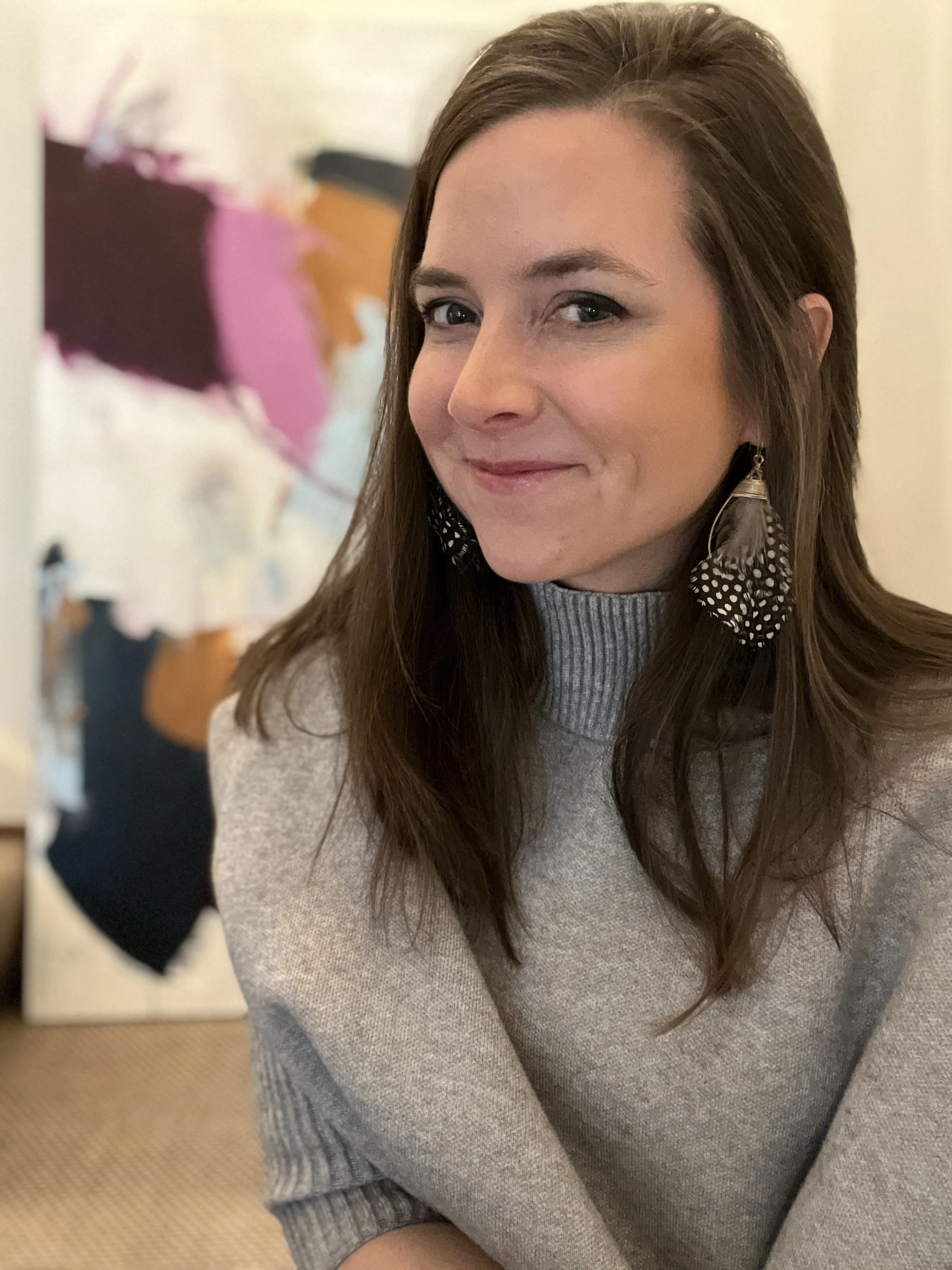 A woman with long brown hair wearing a gray sweater and feather earrings.