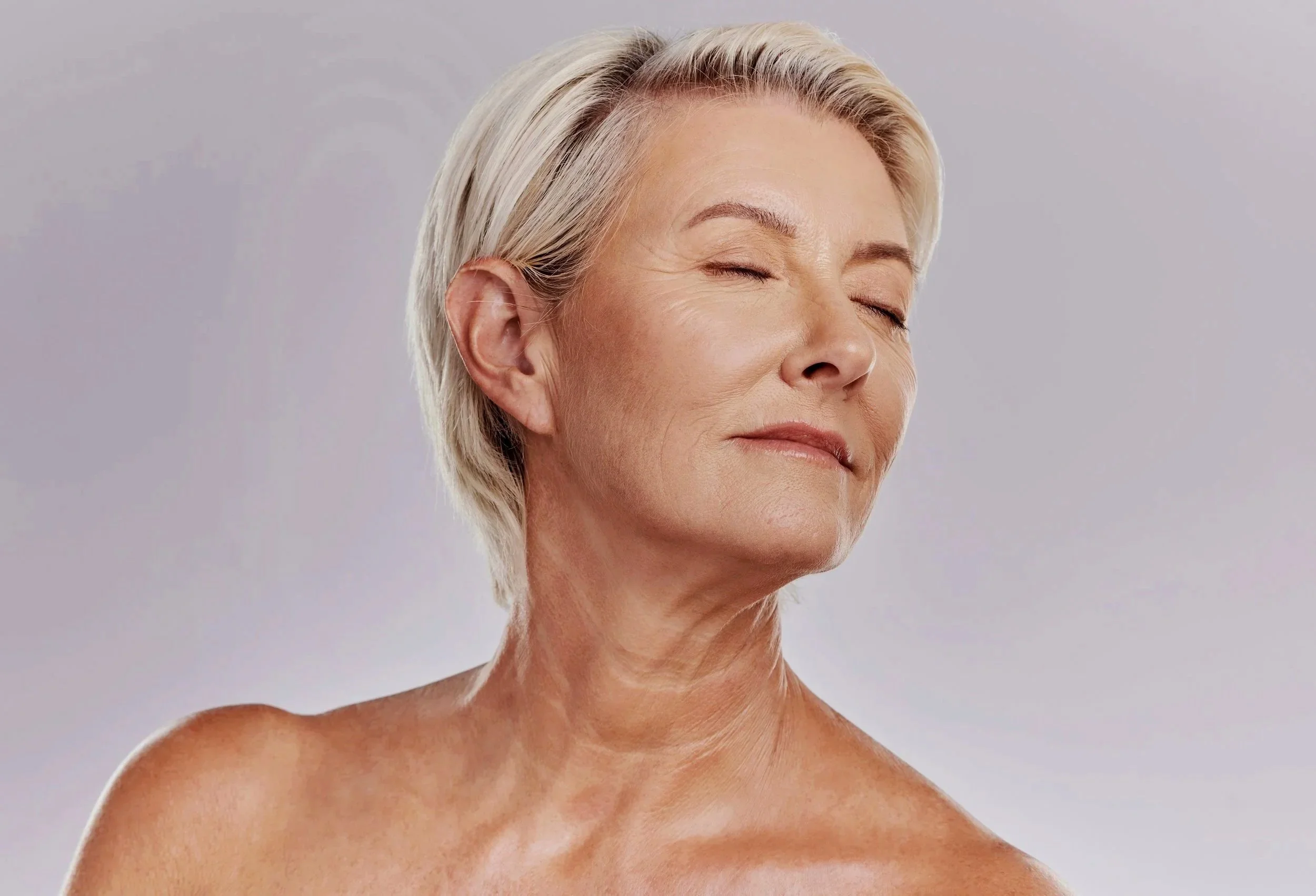 Close-up of an older woman with short, platinum blonde hair, closed eyes, and relaxed expression, against a neutral background.