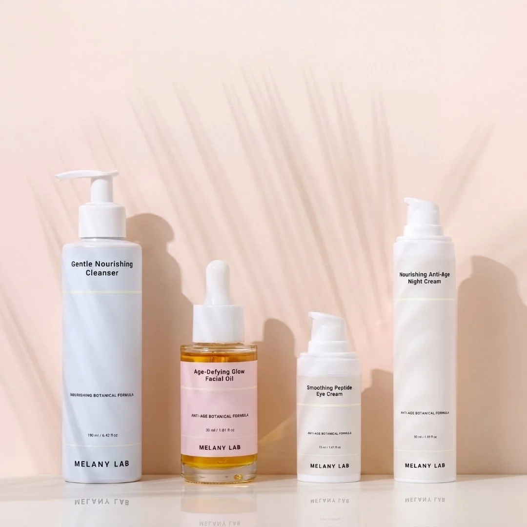 Four skincare products on a white surface against a light pink background: from left, a tall bottle labeled 'Gentle Nourishing Cleanser,' a small bottle labeled 'Age-Defying Glow Facial Oil,' a small pump bottle labeled 'Smoothing Peptide Eye Cream,' and a tall bottle labeled 'Nourishing Anti-Age Night Cream.' Shadows cast by the products are visible.