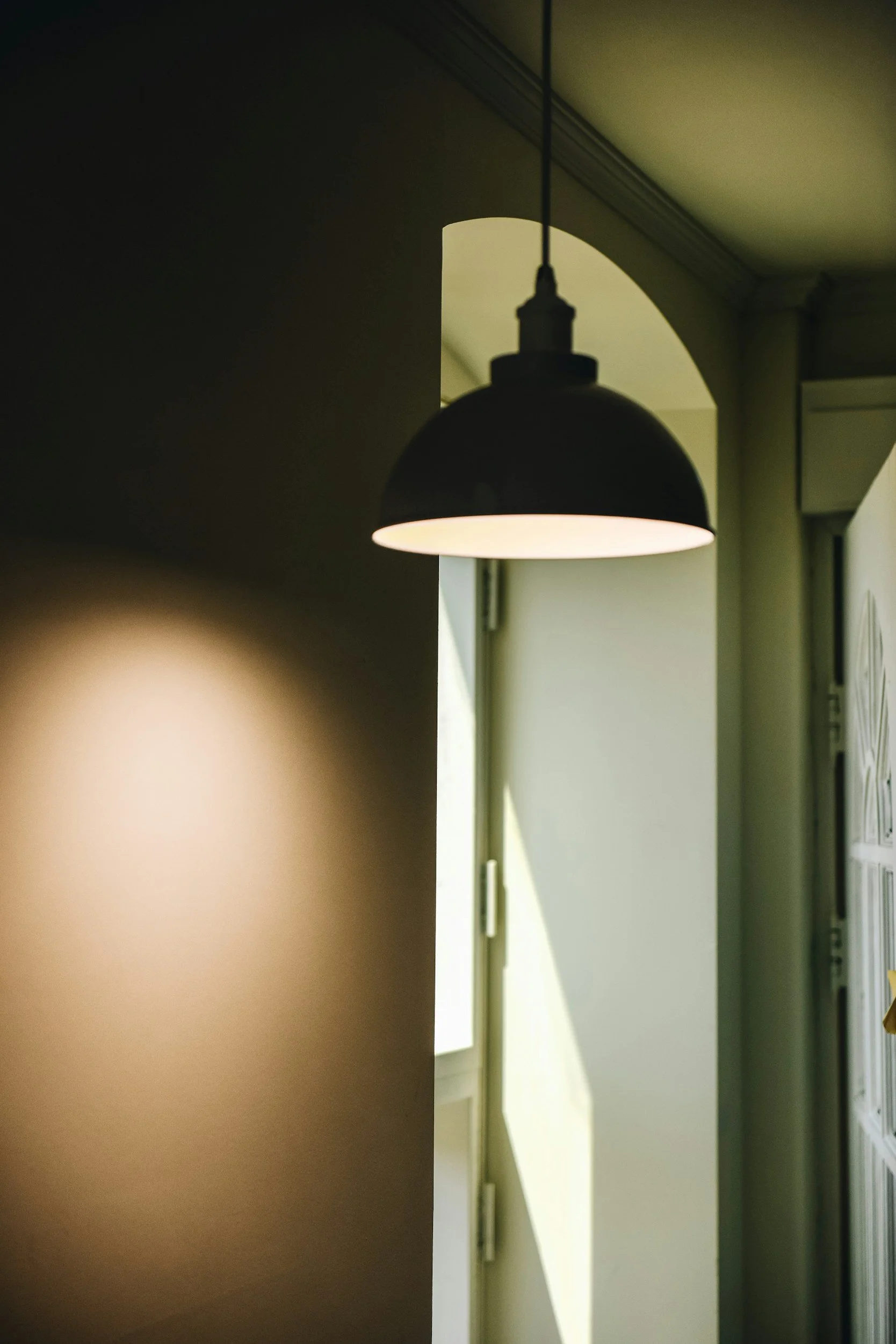 Interior view of a room with a hanging black pendant light, partly illuminated wall, and partly visible doorway or window with natural light in the background.