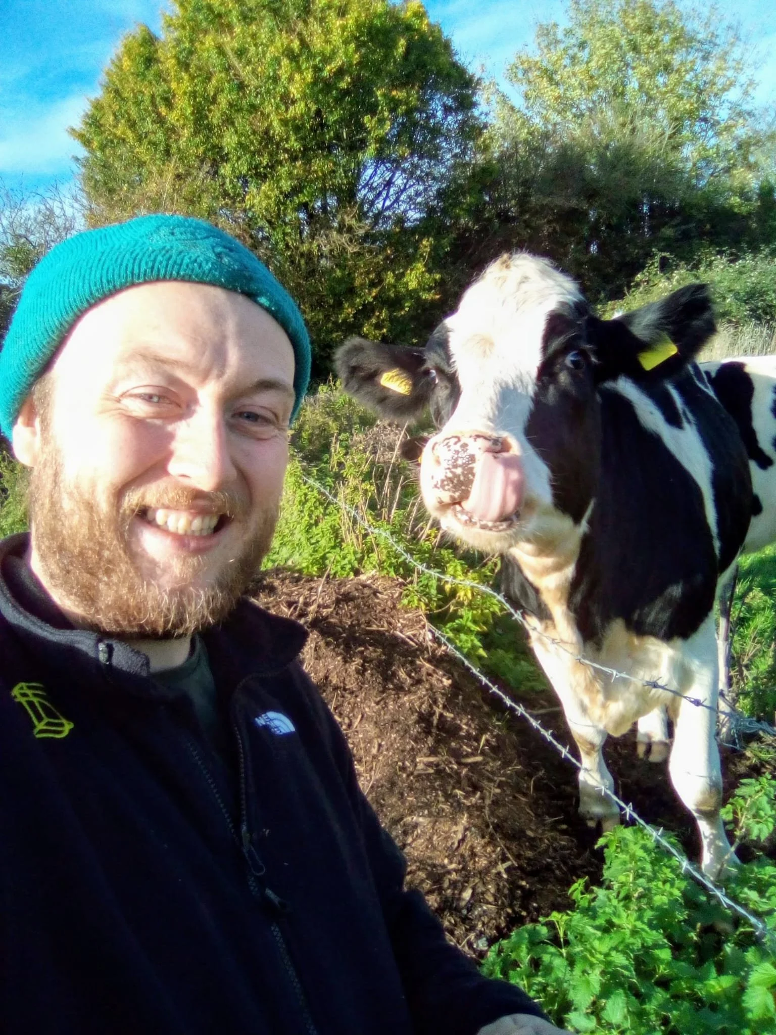 A man smiling outdoors wearing a teal beanie and black jacket, with a black and white calf looking at the camera, surrounded by green foliage and trees.