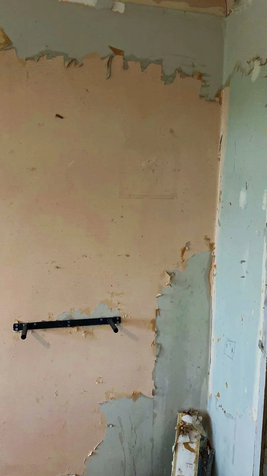 A wall with peeling paint and exposed drywall, with a black horizontal mounting bracket and a partially torn piece of wallpaper or a poster on the floor.