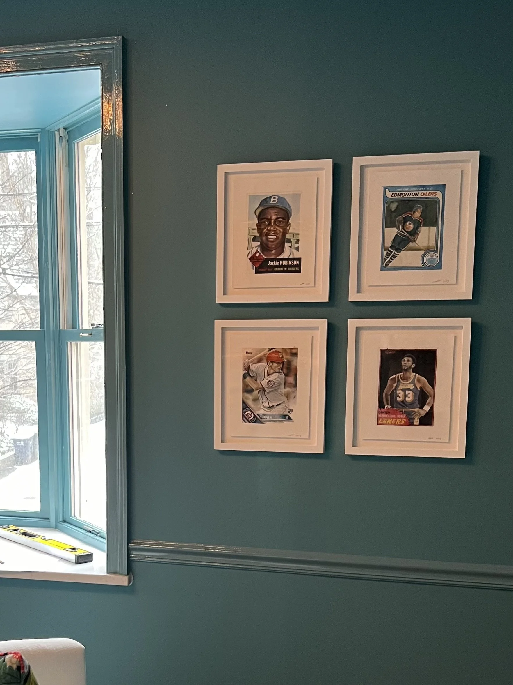 A teal wall with four framed sports posters, three featuring hockey players and one with a basketball player, near a window showing trees outside.