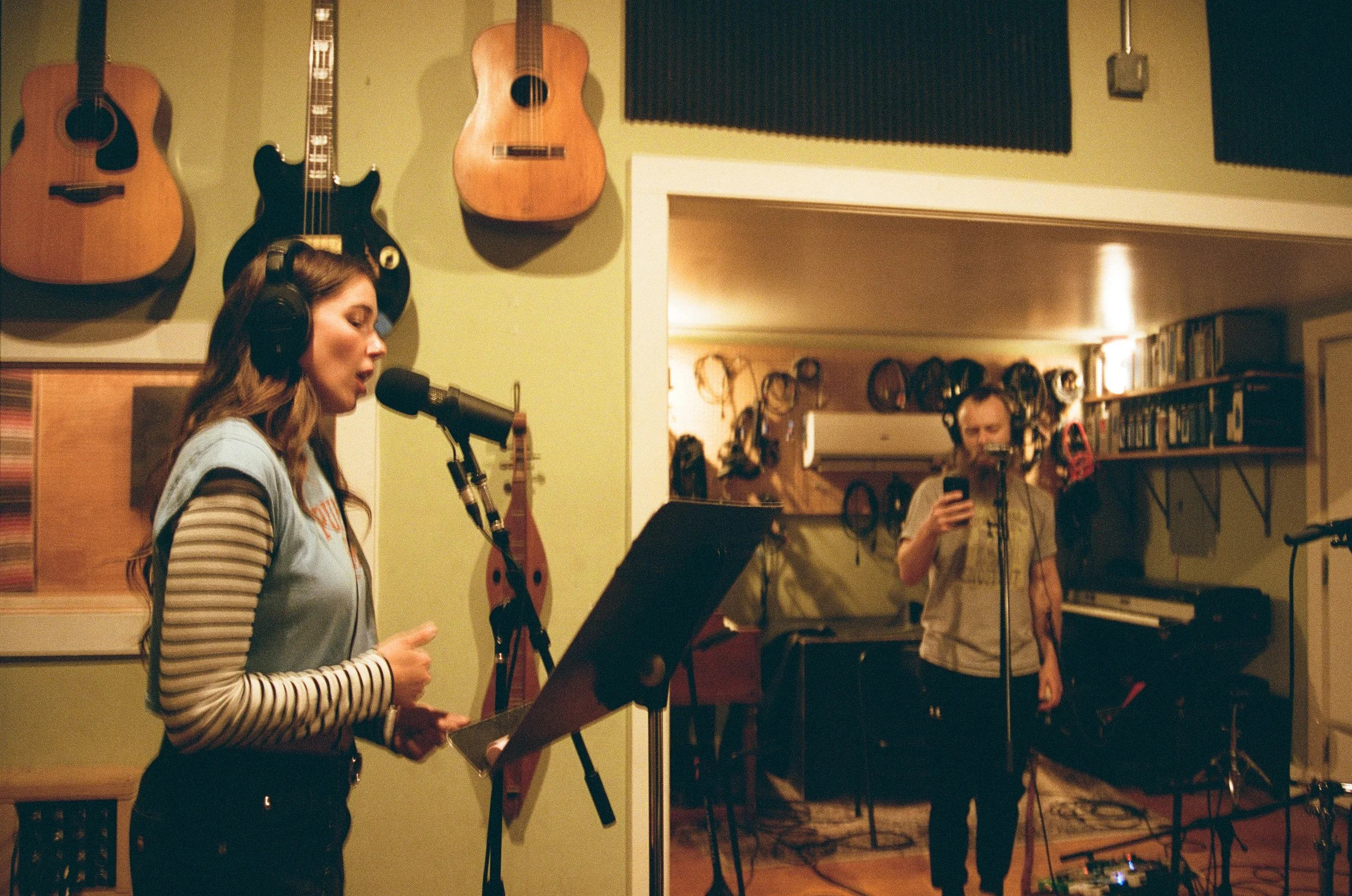 A woman singing into a microphone while playing piano, with a man in the background holding a smartphone in recording mode, in a room decorated with various acoustic guitars hanging on the green wall.