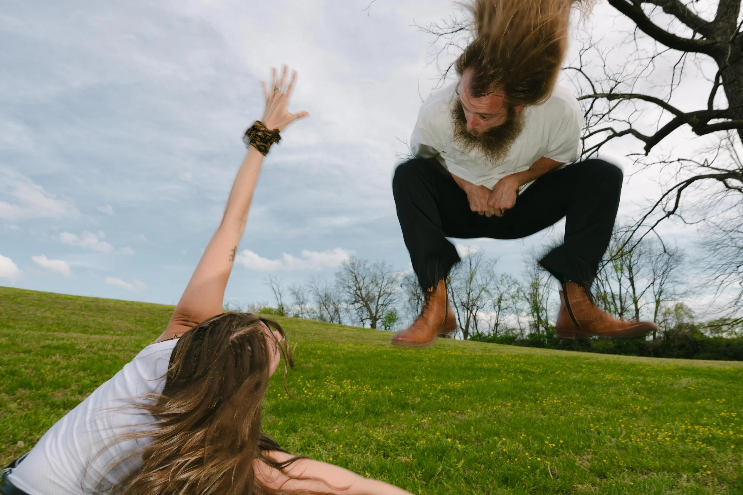 A man with long hair and a beard crouching above a woman lying on the grass, reaching out her hand, as they are outdoors on a cloudy day with trees in the background.