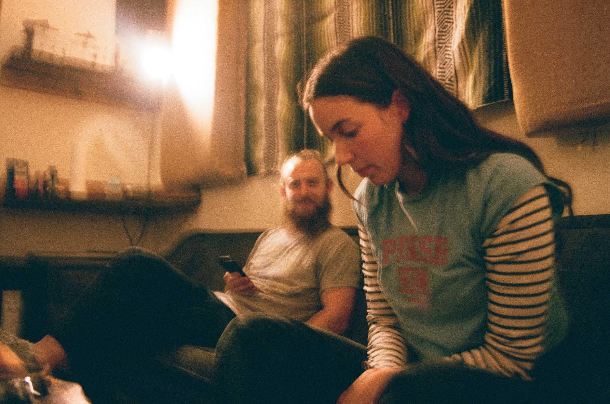 A woman with dark hair looking down and smiling, sitting on a couch in a dimly lit room, with a man with a beard sitting behind her, smiling and looking at her, holding a smartphone. Music being recorded at The Bomb Shelter Studios.
